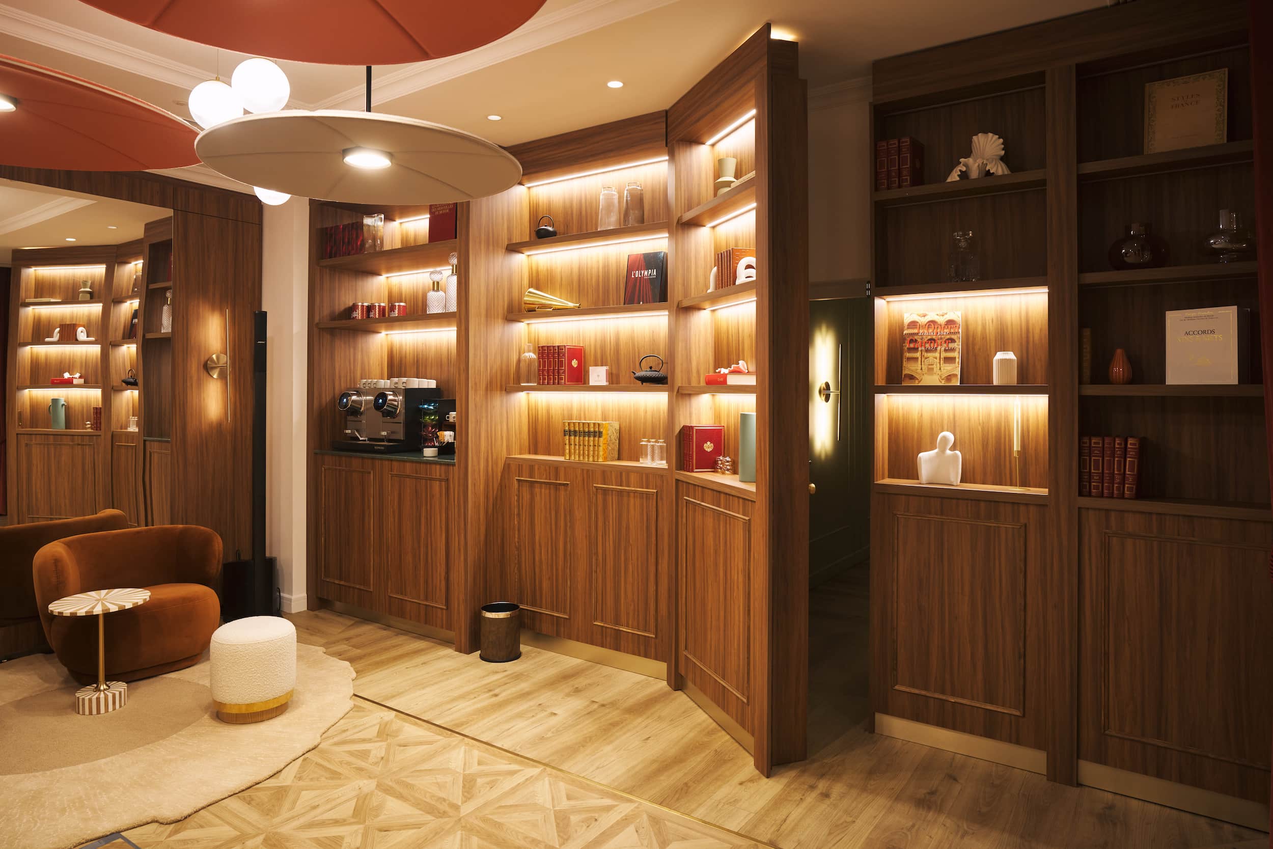 Cozy lounge corner with a brown armchair, white ottoman, small round table, and illuminated wooden shelves holding books and decorative items.