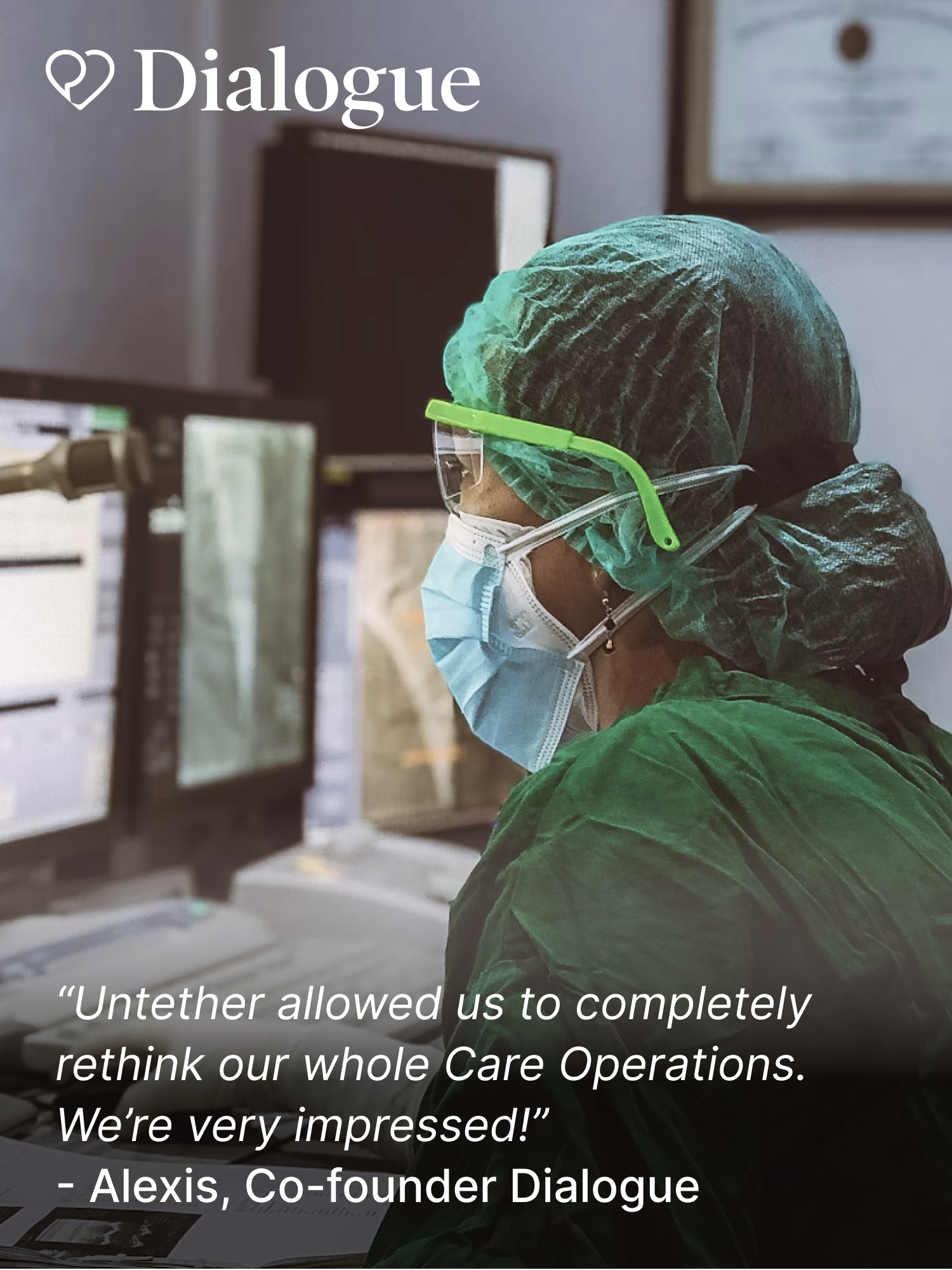 Healthcare worker in green protective gear, face mask, and goggles, monitoring computer screens in a clinical setting.