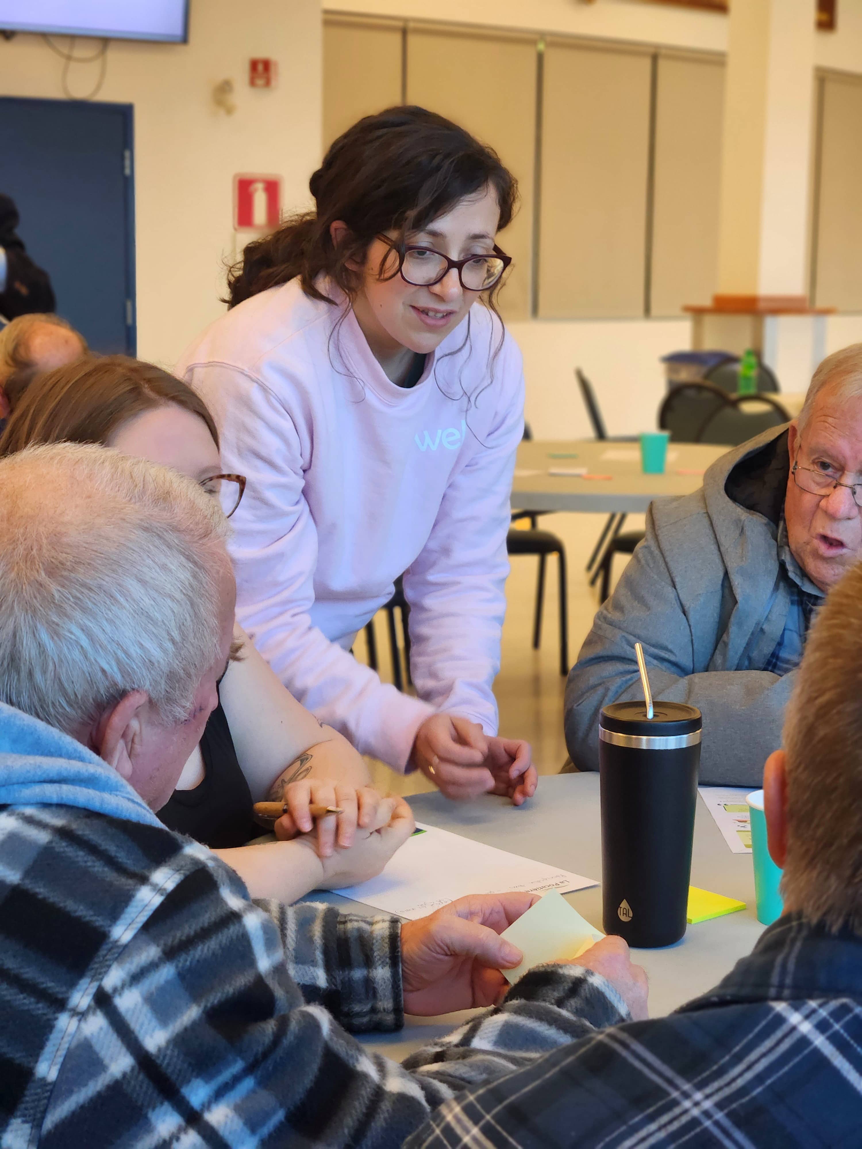 Une animatrice de table explique un atelier aux citoyens assis à une table ronde lors d'une consultation citoyenne.
