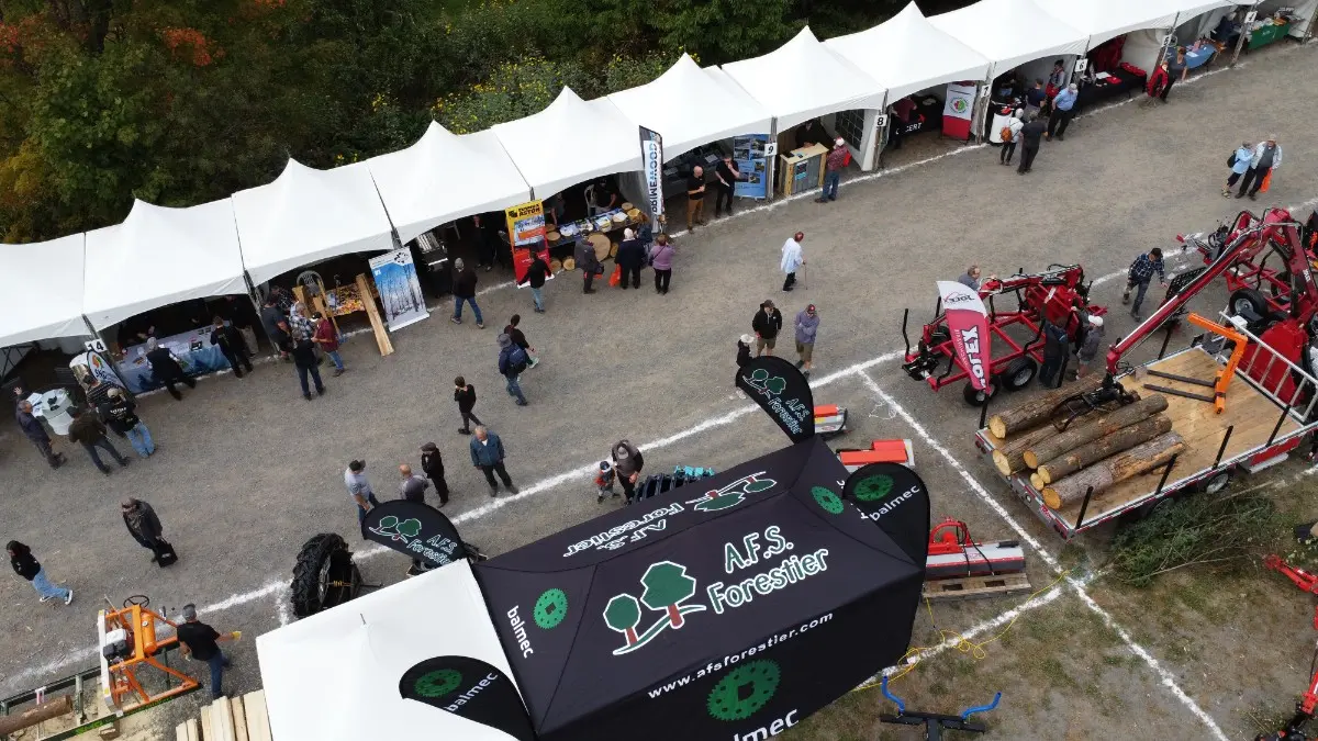 Un homme portant une casquette bleue et un chandail vert utilise un outil traditionnel pour fendre une bûche posée sur un billot, en plein air, lors d’un événement ou d’un atelier de bois. On voit ses mains appuyer fermement sur l’outil pendant qu’il travaille, avec des tentes et des arbres en arrière-plan sous un soleil éclatant.