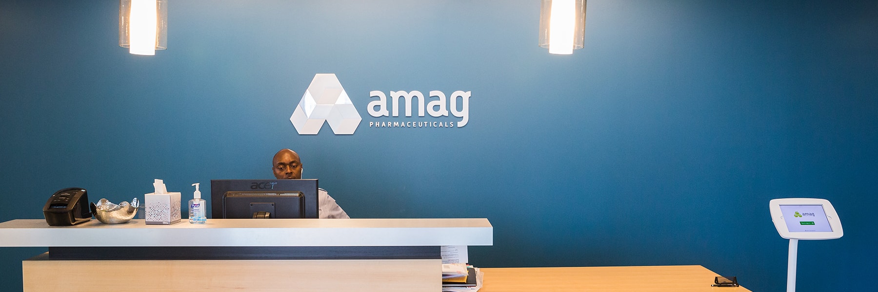 Reception desk with a man working on a computer under the amag Pharmaceuticals logo on a blue wall.