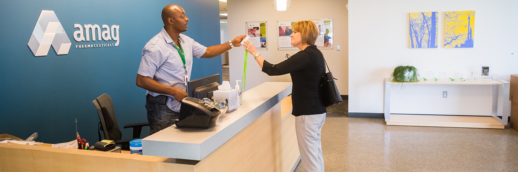 A person behind the amag front desk handing a visitor badge to another person.
