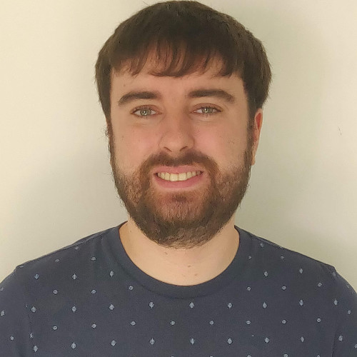 Man with short brown hair and beard smiling, wearing a dark blue shirt with small light patterns.