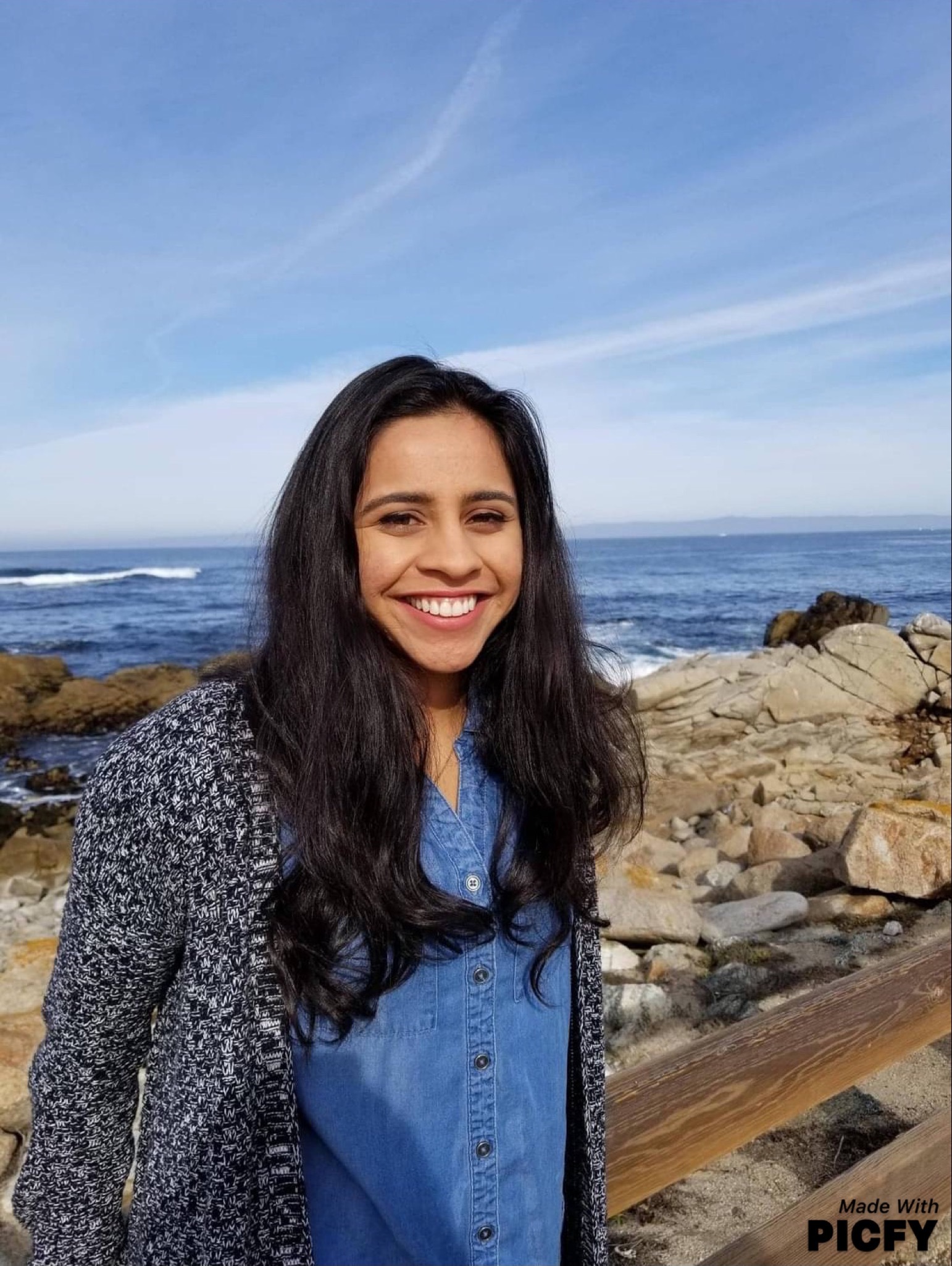 Smiling woman with long dark hair in a blue shirt and gray sweater by rocky ocean shore under blue sky.