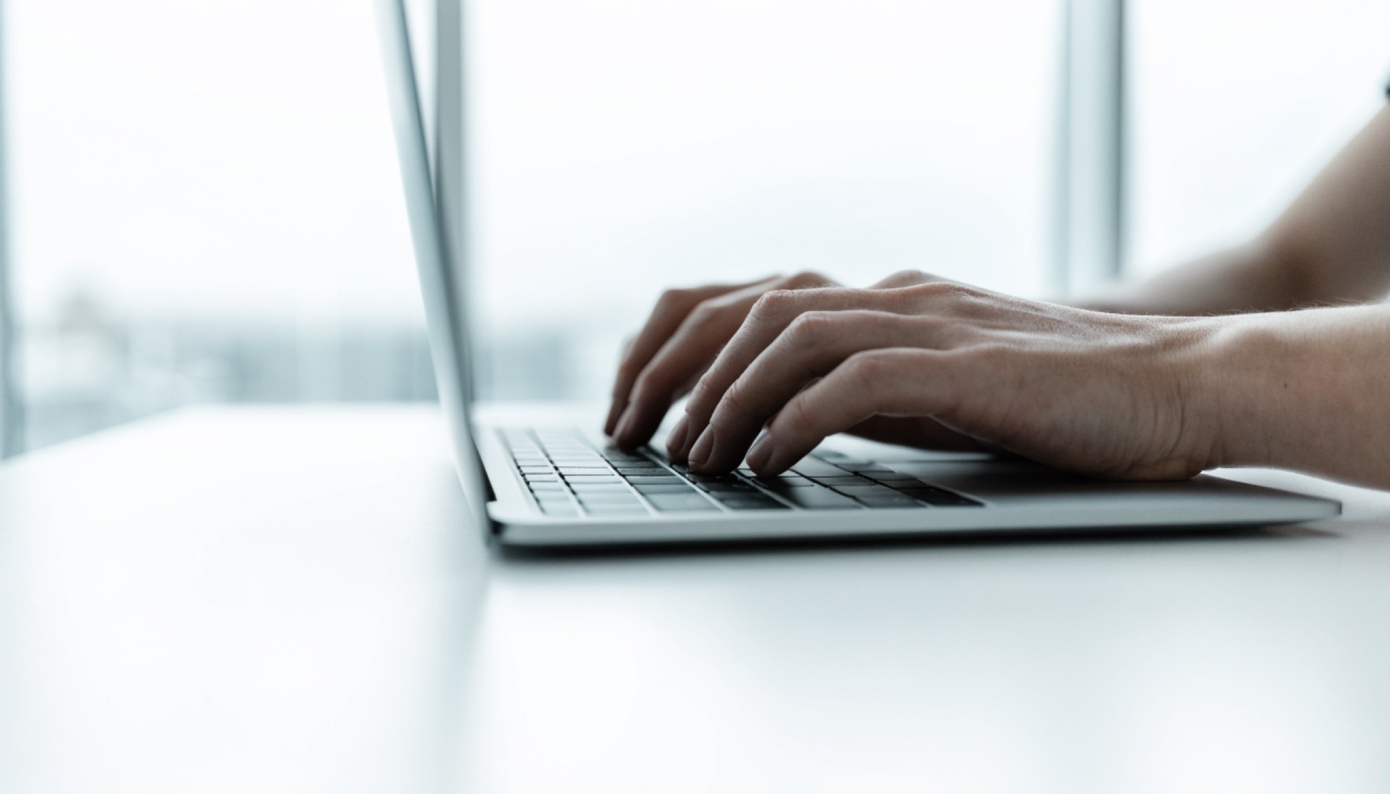 Manos escribiendo en el teclado de un computador portátil sobre un escritorio blanco junto a una ventana