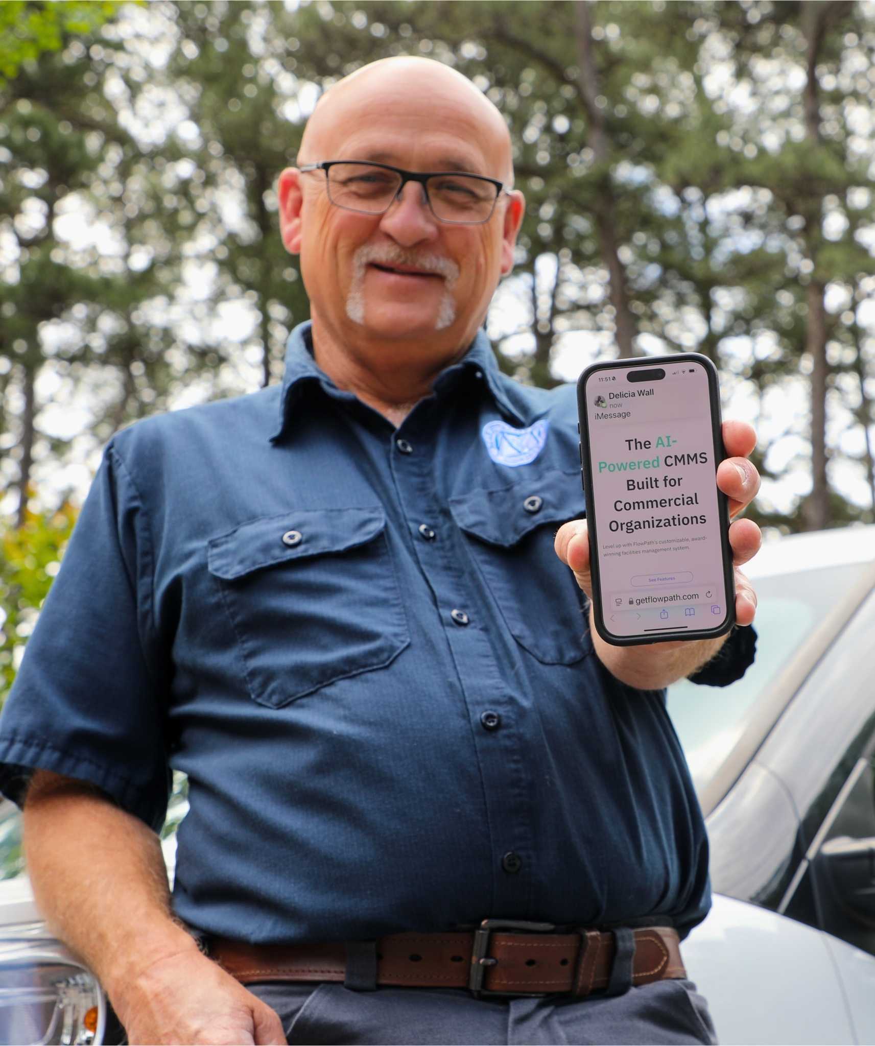 A maintenance worker holds up a phone displaying FlowPath CMMS, built for schools, commercial organizations, municipalities, and more.