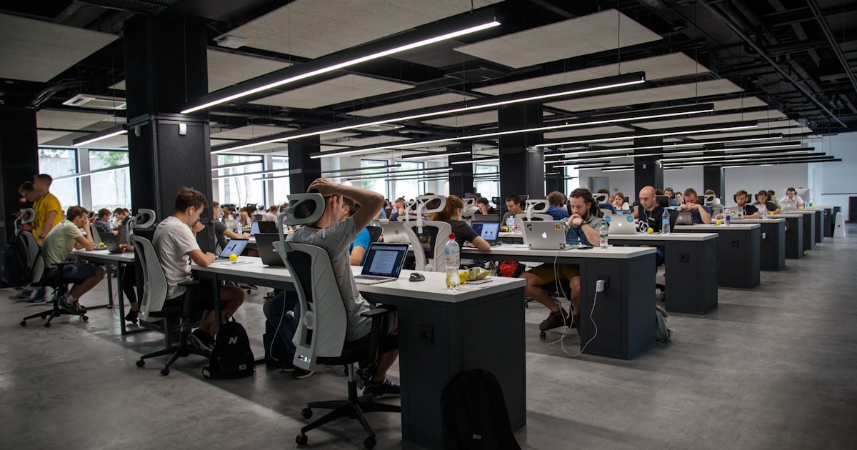 Open office workspace with scattered notes and multiple screens representing disconnected maintenance tracking systems