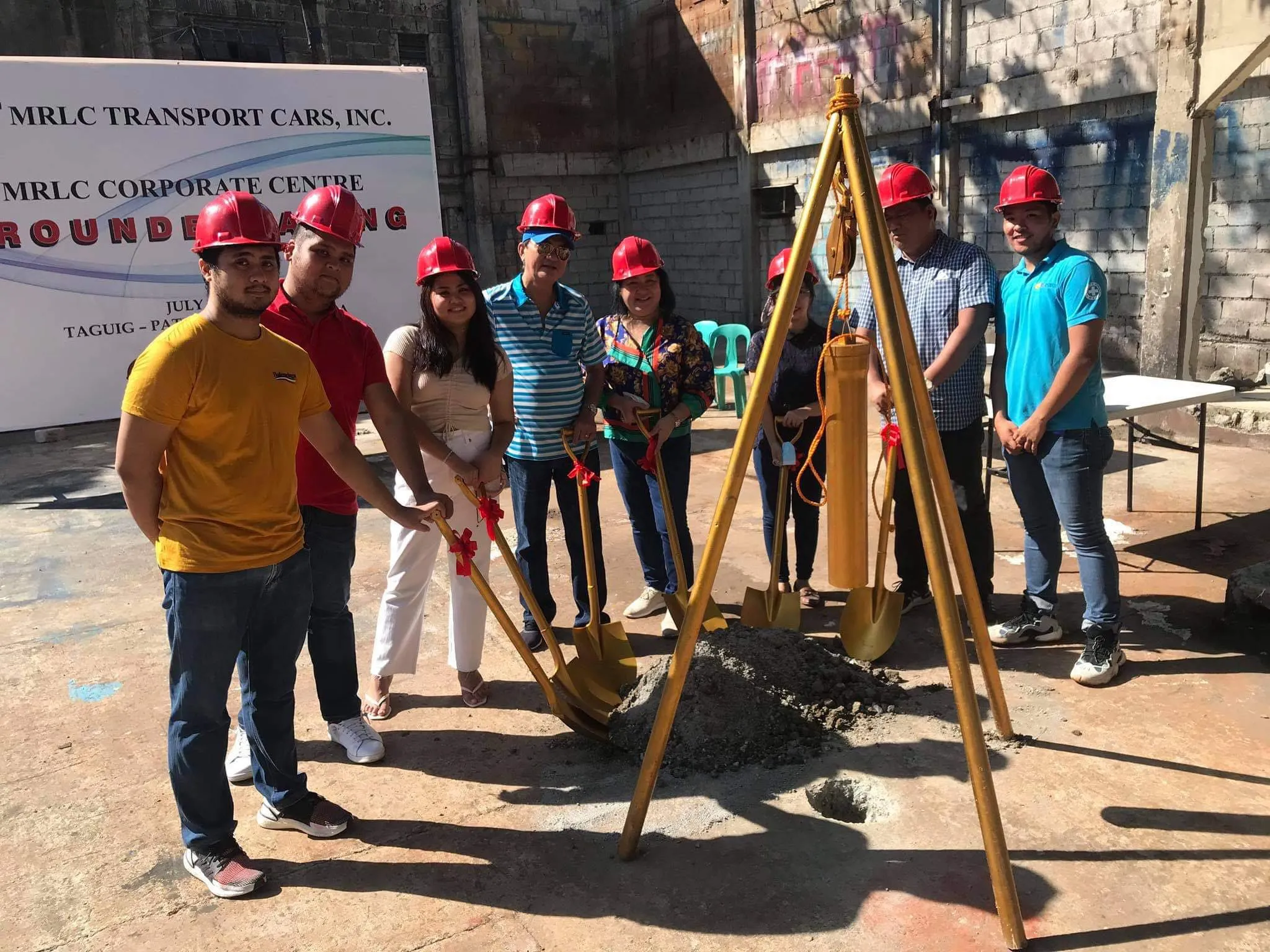 Group of people wearing red hard hats holding shovels at a groundbreaking ceremony.