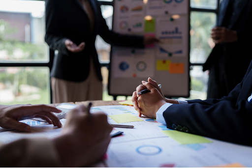 Business professionals in a meeting with documents and charts. Hands gesture over papers in foreground, while someone presents at a whiteboard with diagrams in the background