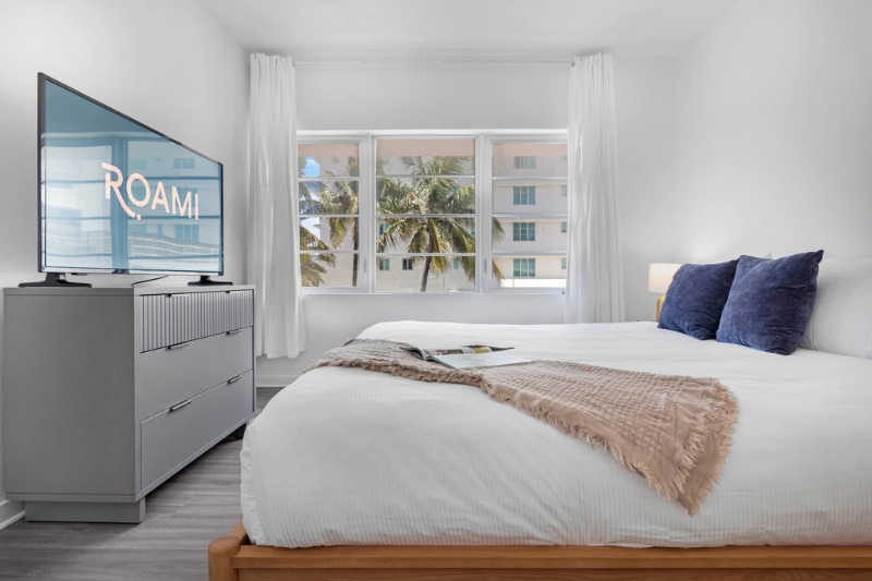 white bedroom with navy pillows tv and window view of palm trees at 1200 ocean by roami