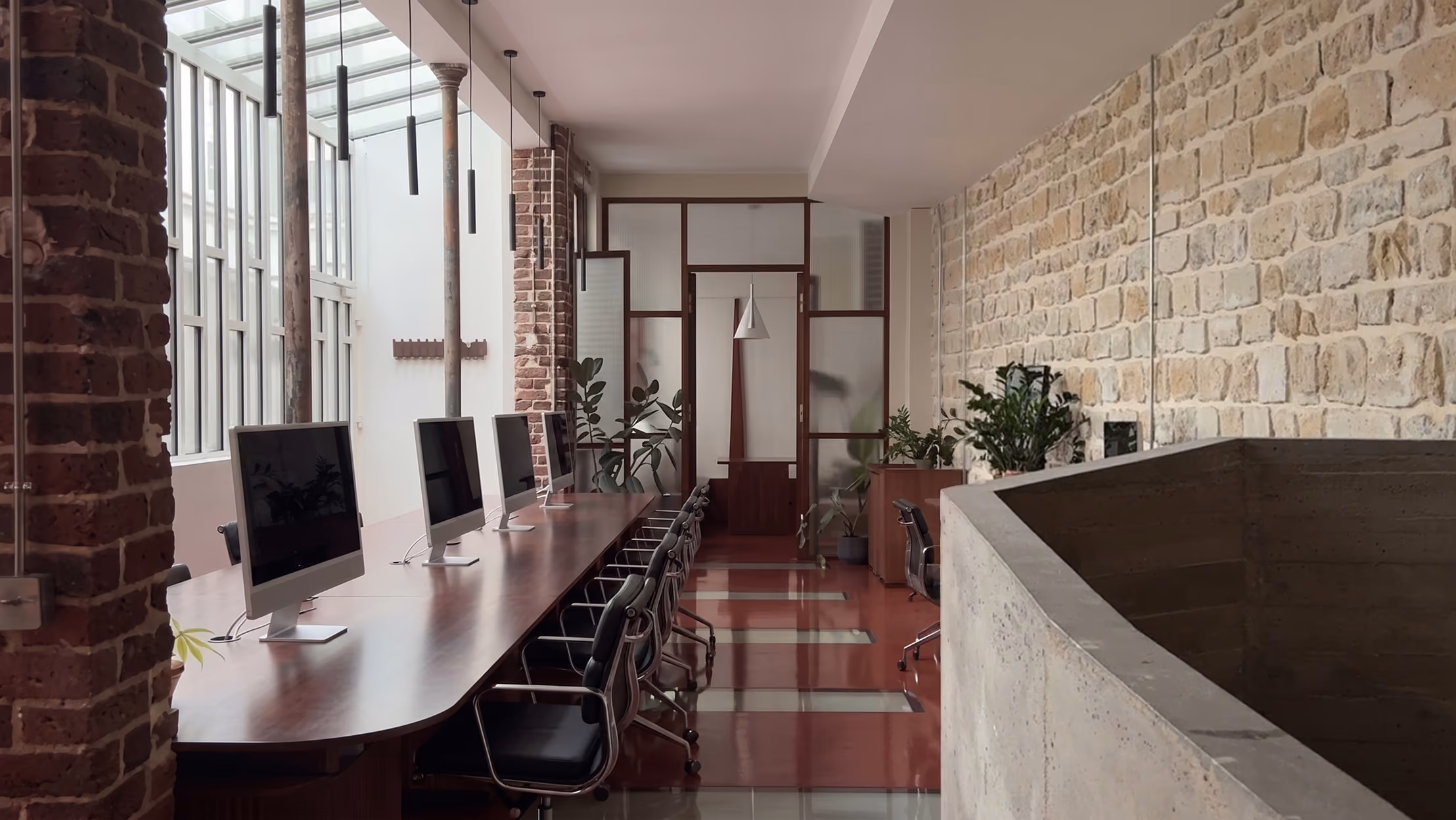 Modern office space with a long wooden desk holding five desktop computers and black swivel chairs, set against large windows and exposed brick and stone walls with hanging pendant lights and potted plants.