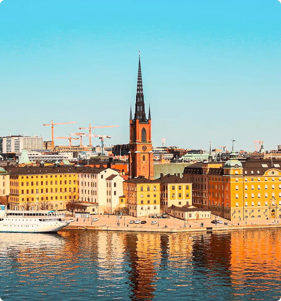 Waterfront view of colorful buildings and a tall church spire reflected in the water under a clear blue sky.