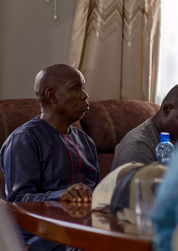 Two men sitting at a wooden table, one wearing a dark blue traditional outfit with red embroidery, in a room with beige curtains and a brown couch.