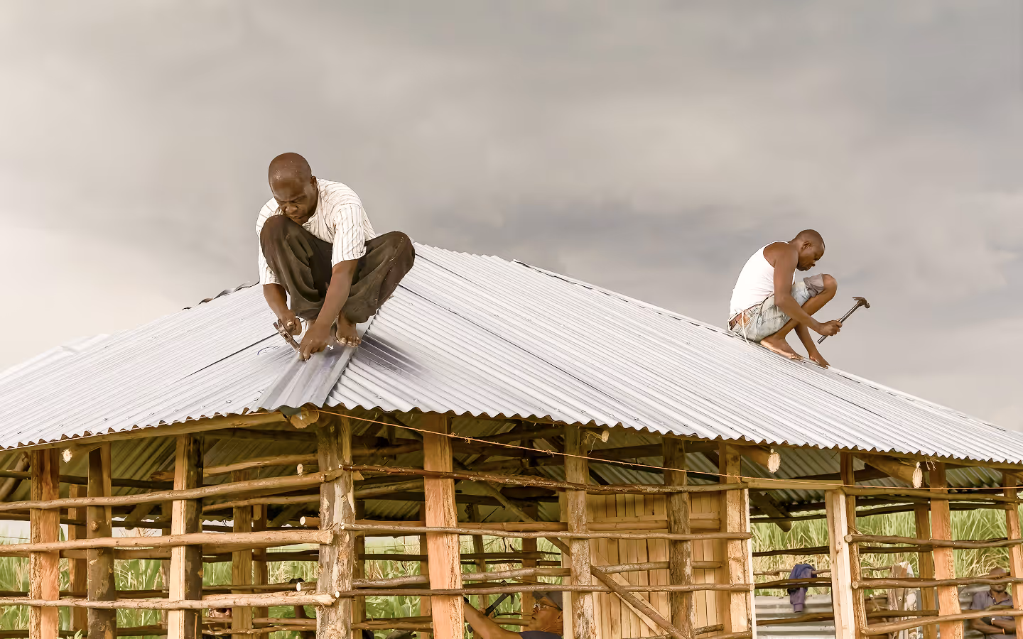 Two men working barefoot to install a corrugated metal roof on a wooden structure under a cloudy sky.