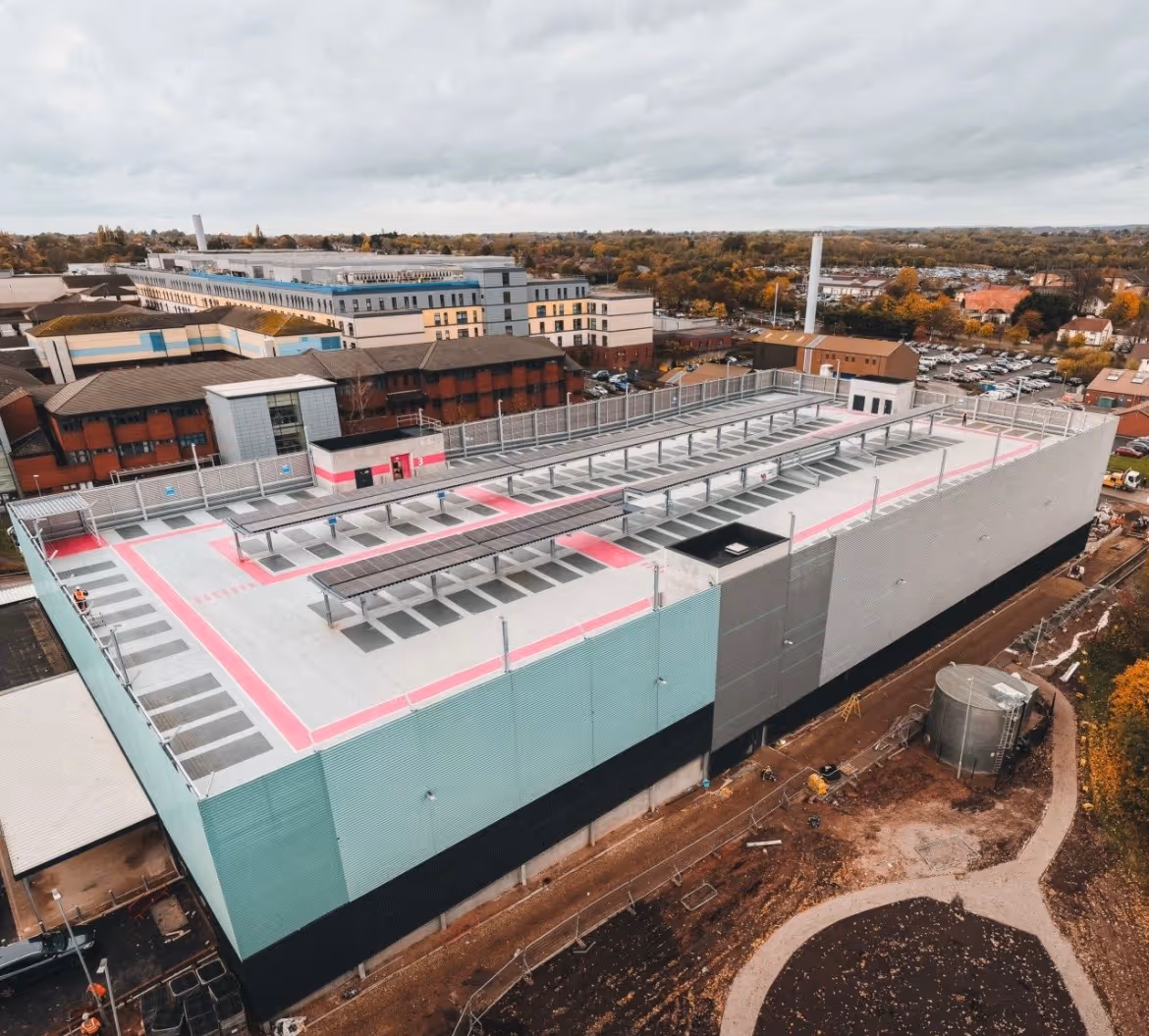 Aerial view of a large multi-story parking garage with solar panels installed on the roof and a surrounding urban area with buildings and trees.