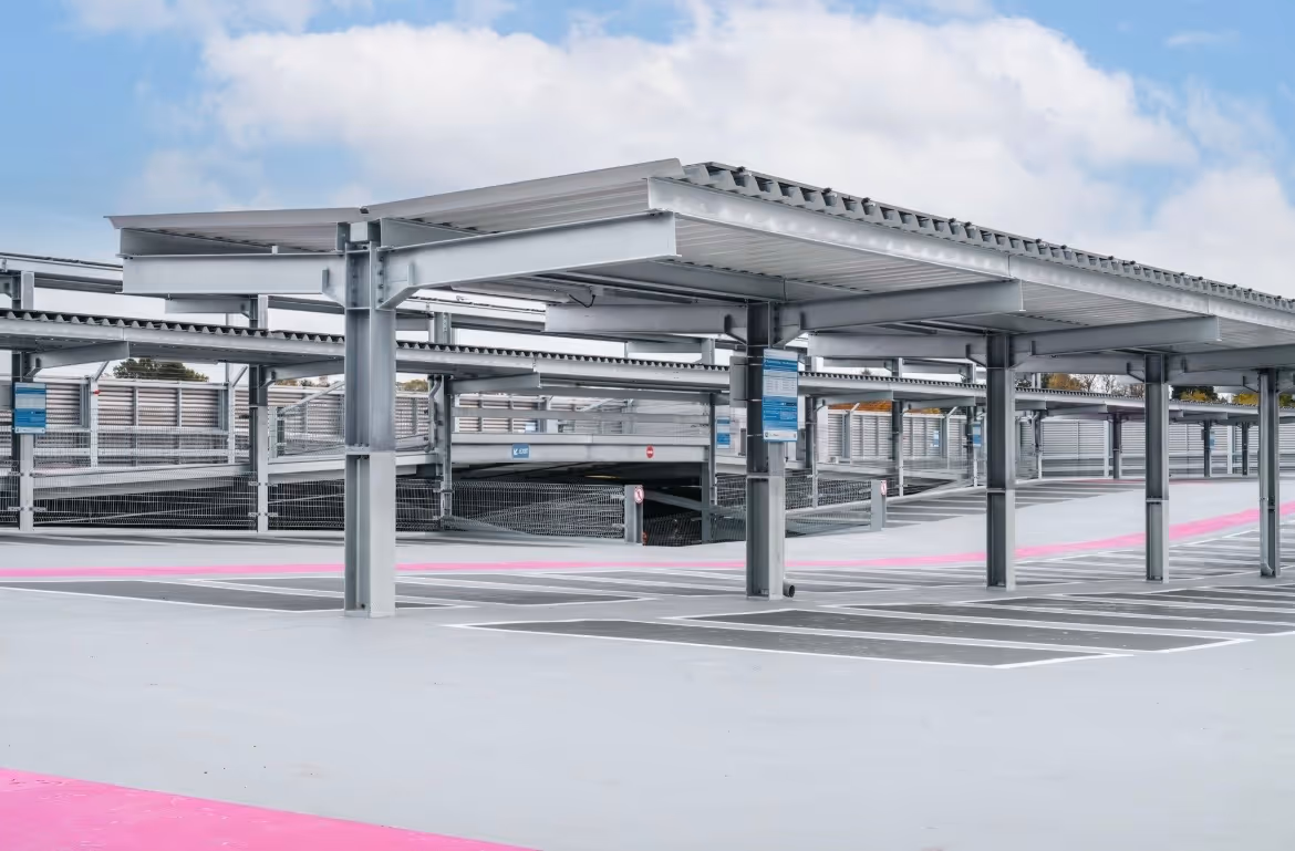 Empty outdoor parking lot with metal canopy structures and marked parking spaces under a partly cloudy sky.