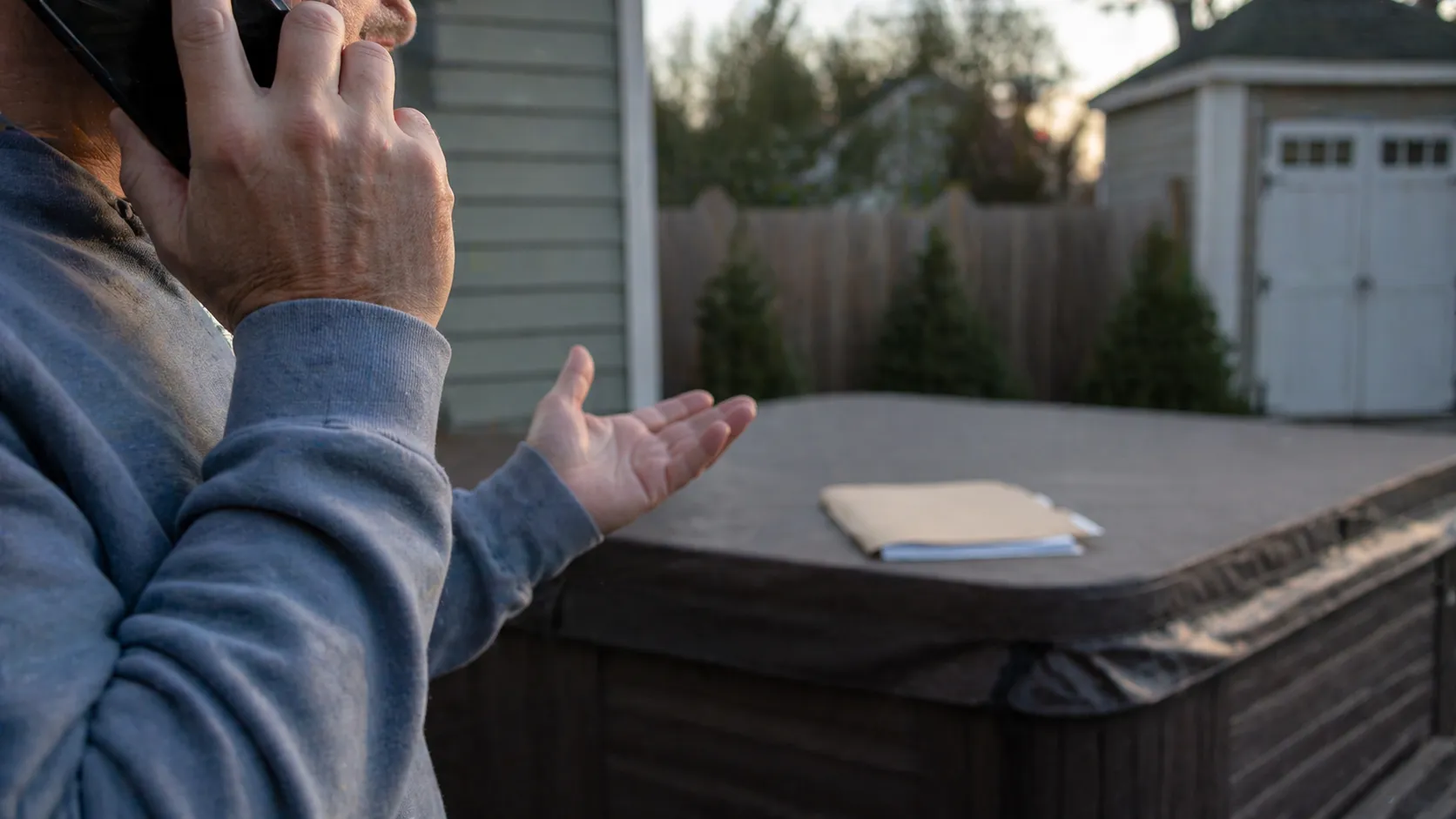 Close-up of a person on the phone beside a covered hot tub in a backyard with paperwork resting on top, suggesting service or warranty frustration