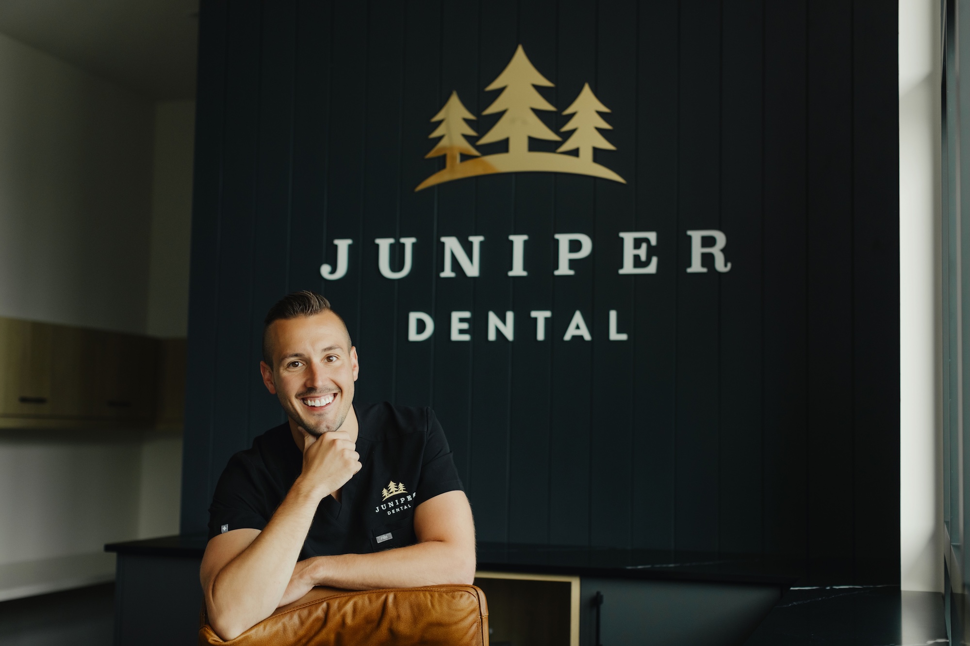 Dr. Chris Cantlon at his front desk at Juniper Dental, the logo with three trees is behind him on a dark green wall.