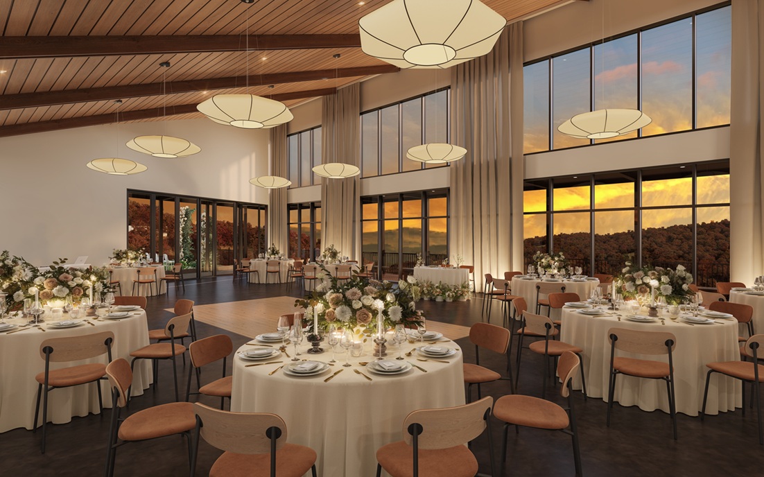 Elegant banquet hall with round tables set with white tablecloths, floral centerpieces, and brown chairs under wooden ceiling with modern pendant lights at sunset.