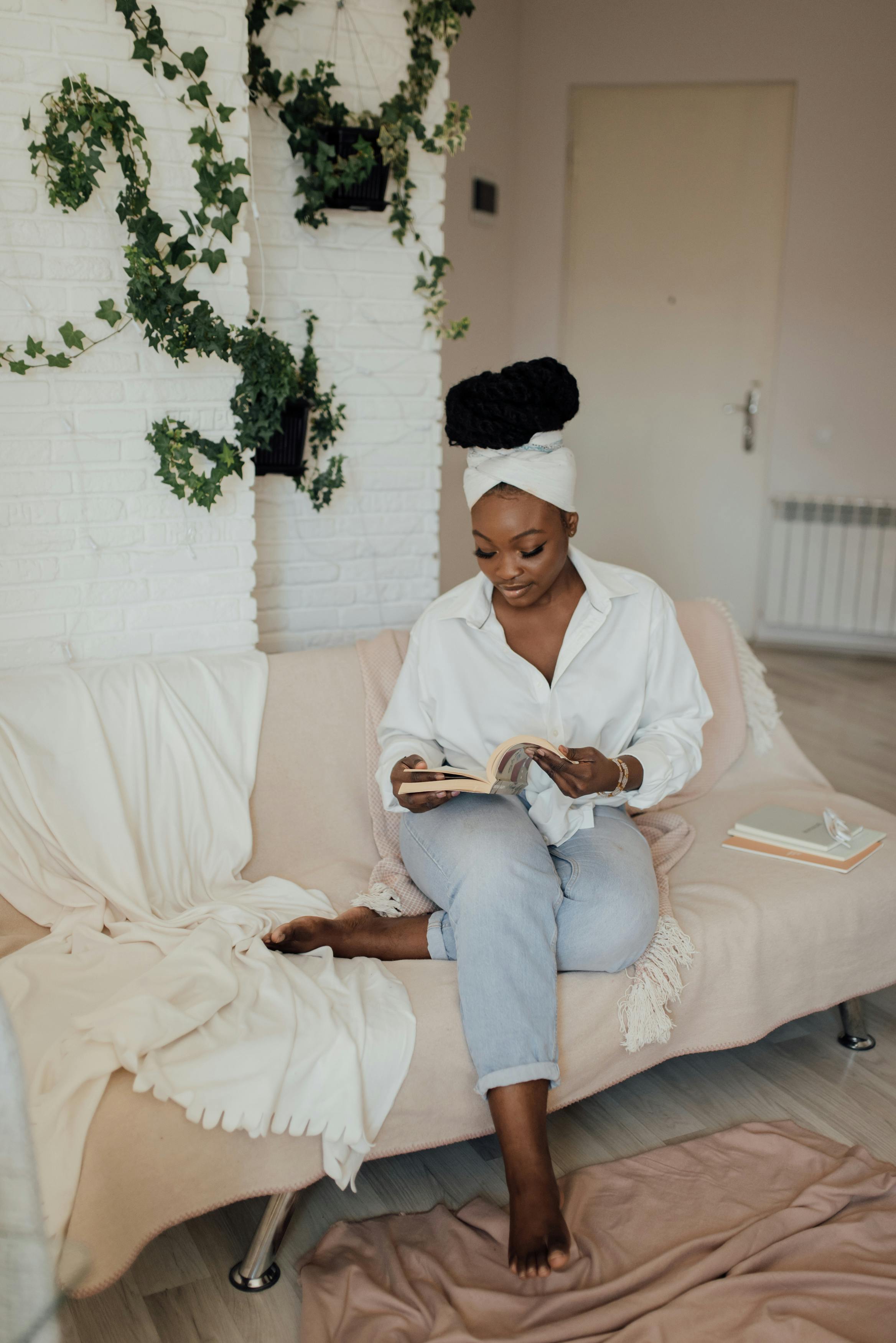 A woman sitting peacefully on a neutral-toned sofa, reading a book. She is wearing a white button-down shirt and a head wrap, surrounded by minimalist home decor and climbing ivy.