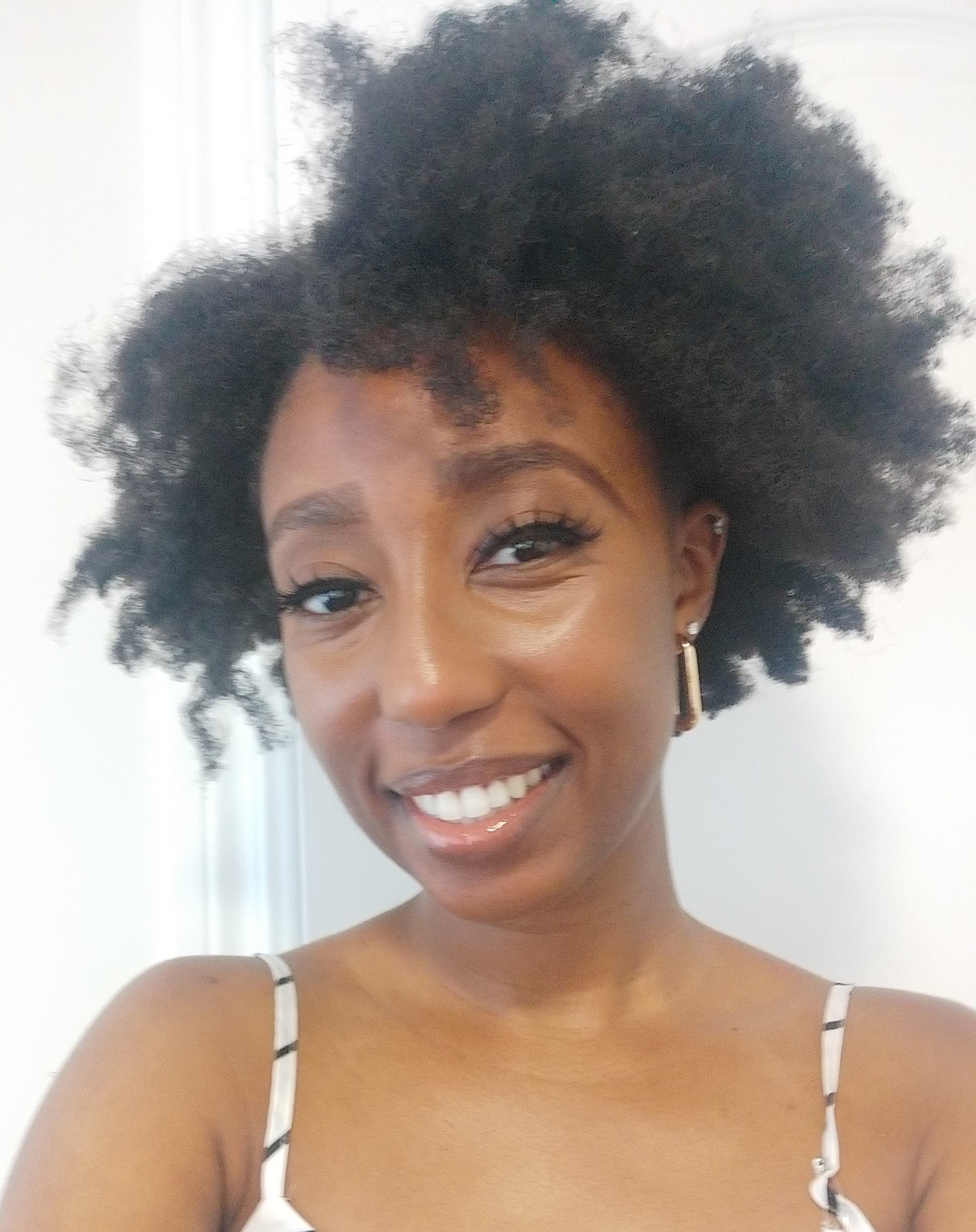 A smiling portrait of SH, founder of Jasmine Anthea, showcasing her natural hair texture against a clean, bright background. She is wearing gold earrings and a minimalist white and black patterned top.