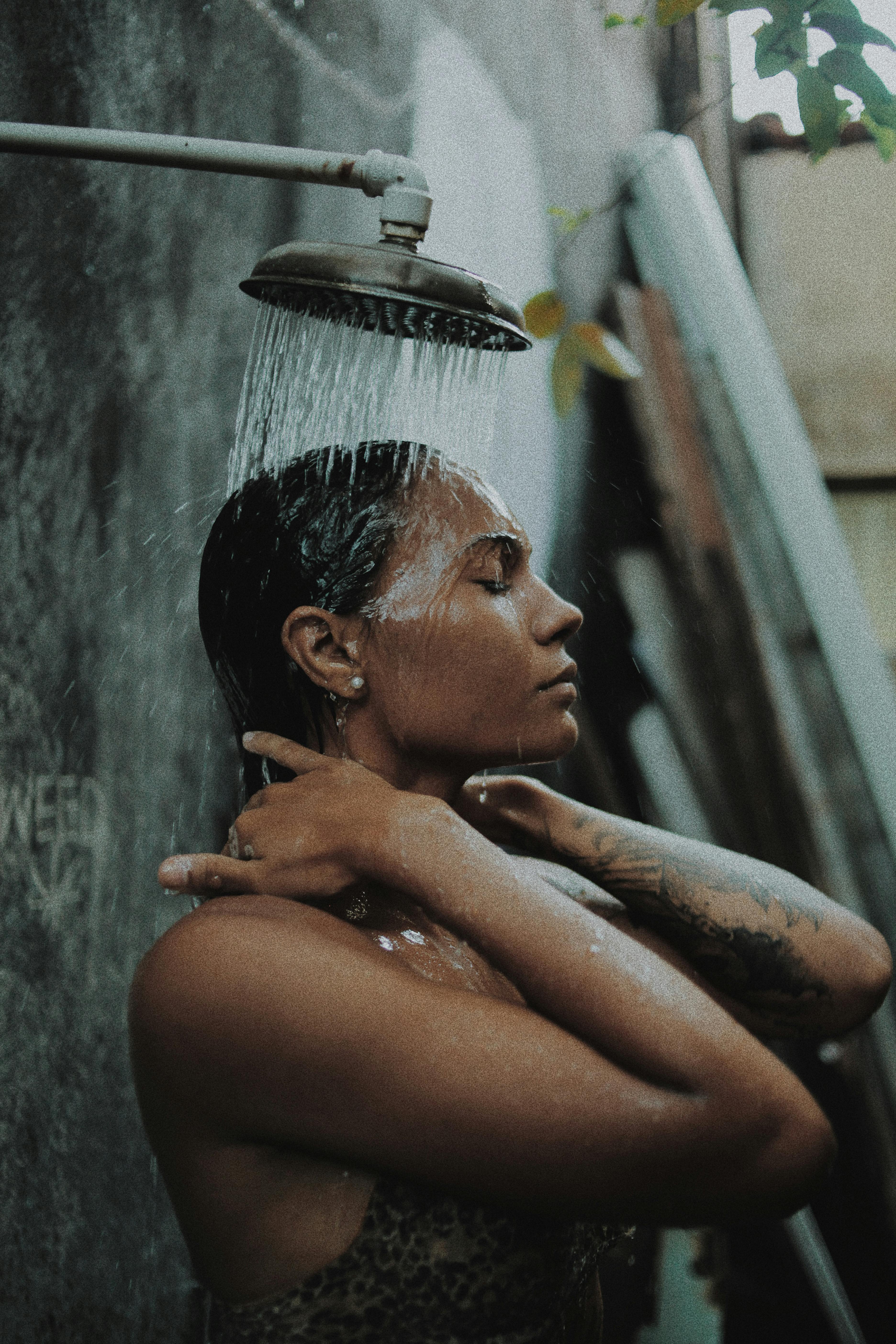 A profile view of a woman with her eyes closed, standing under a rustic metal shower head as water pours over her hair and shoulders against a weathered wall.