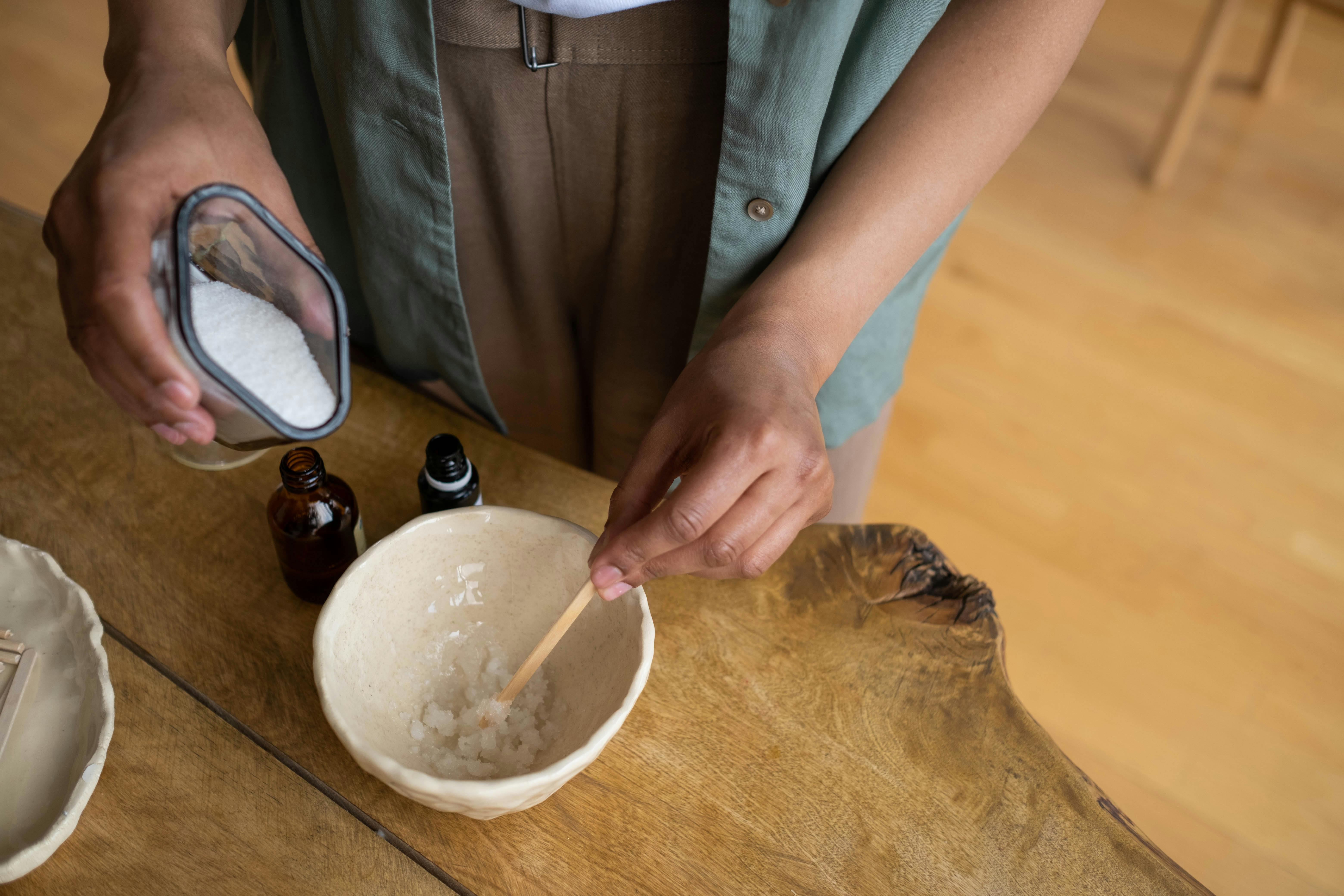 An overhead shot of hands pouring white salts from a glass container into a ceramic bowl, with small dark apothecary bottles resting on a wooden table.