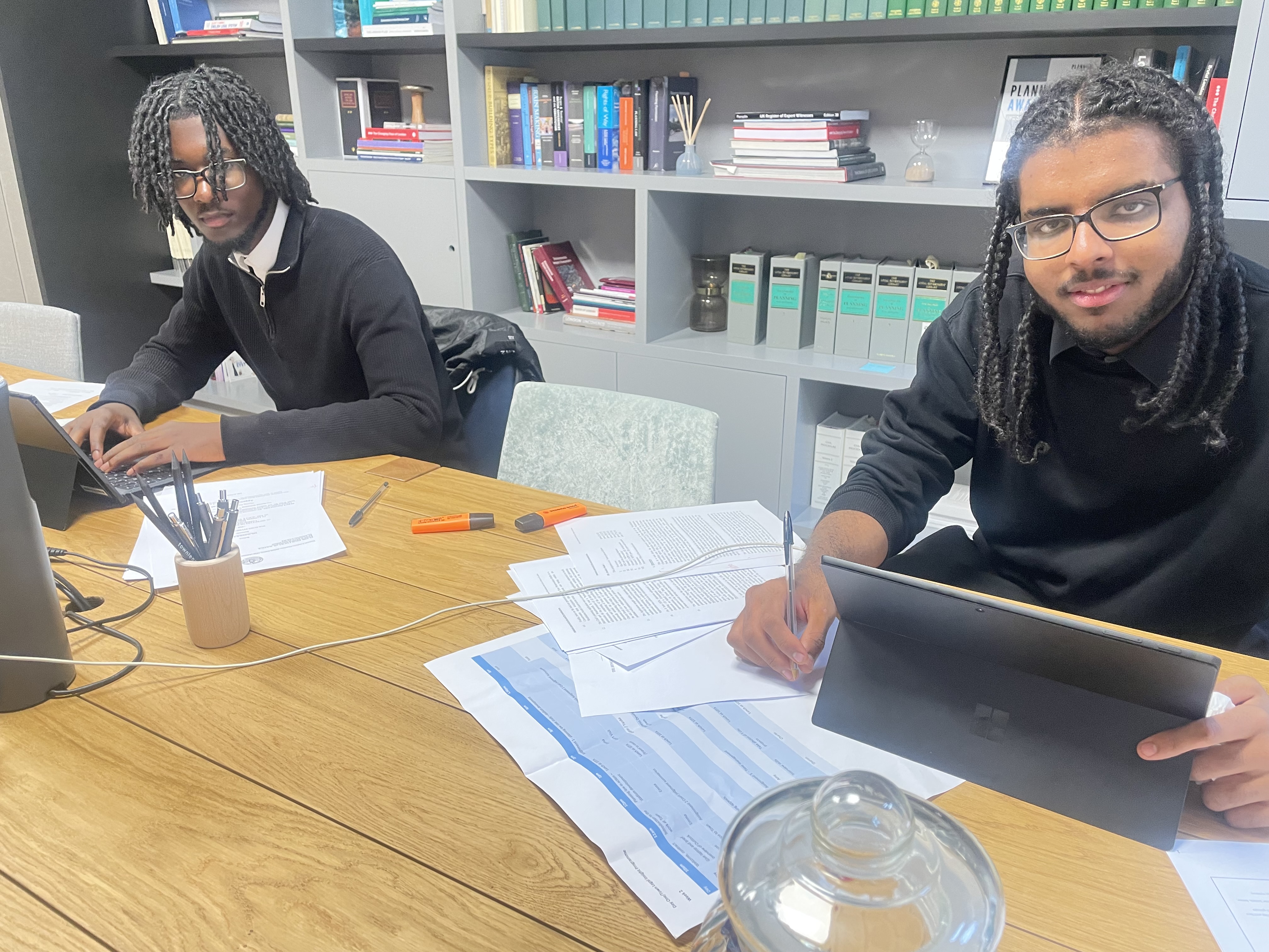 Reuben and Sudi sitting in an office at a large desk with paper, pens and laptops. They are dressed in smart black clothes, looking at the camera as they work.