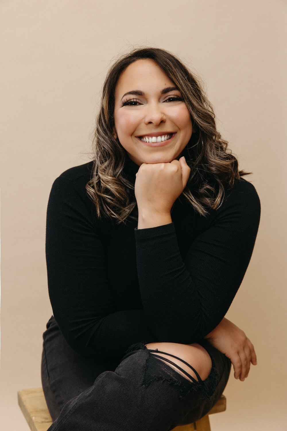 Film director Taty Ortiz wearing a black long-sleeve top and ripped black jeans sitting on a wooden stool against a beige background.