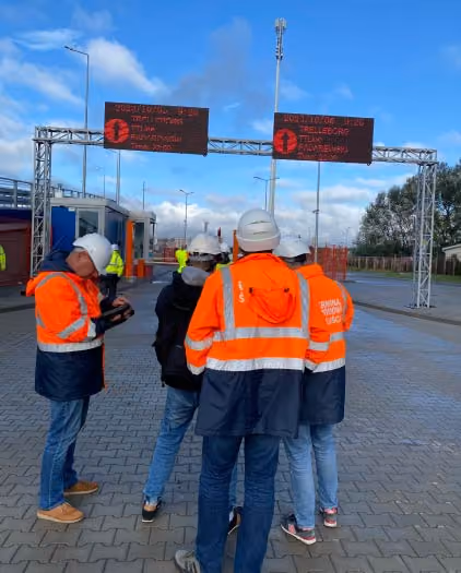 Grupa czterech pracowników budowlanych w pomarańczowych kamizelkach i białych kaskach stoi na utwardzonym terenie pod dwoma cyfrowymi znakami drogowymi.
