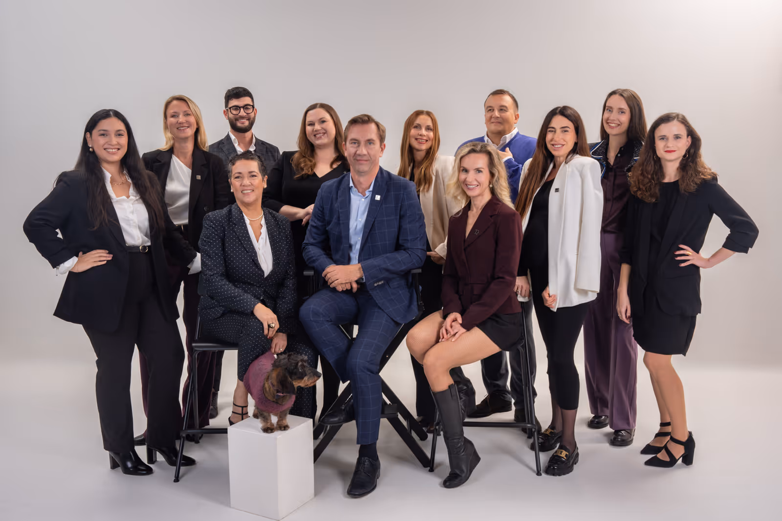 Group of professionally dressed men and women posing together in a studio with a small dog wearing a sweater sitting on a white pedestal.