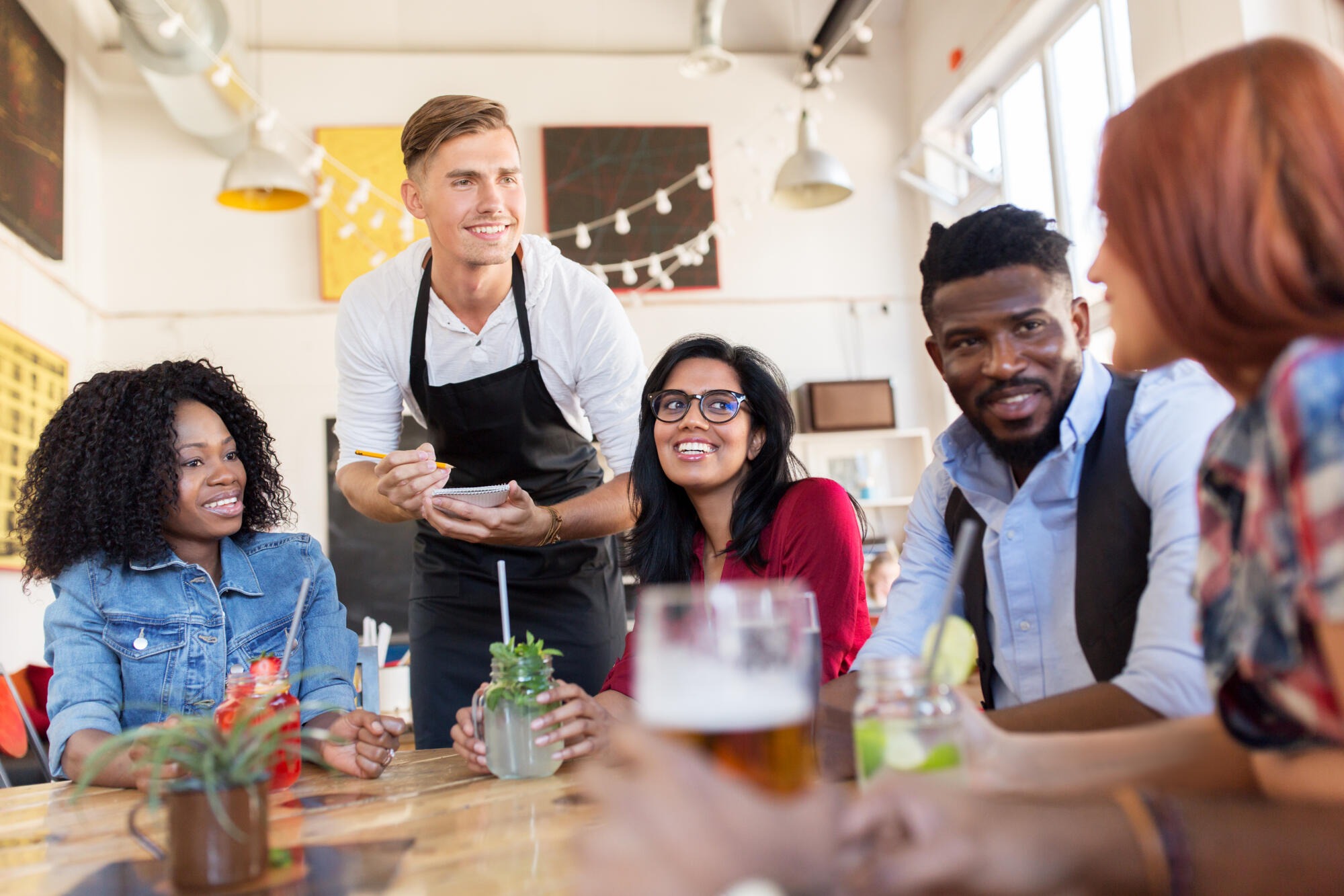 group of happy international friends with drinks and waiter with notepad receiving order at bar or restaurant
