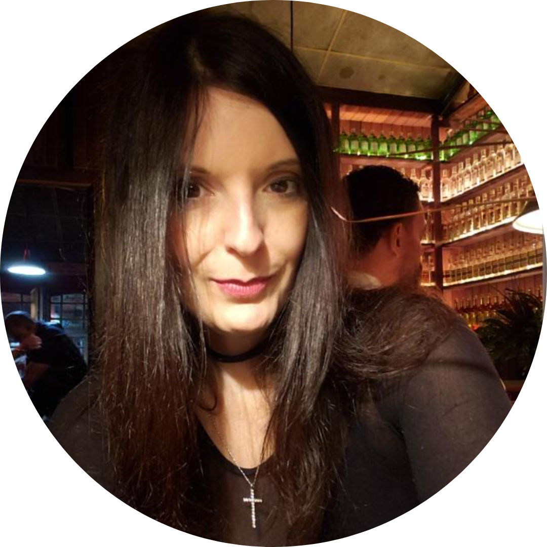 Woman with long dark hair wearing a black top and a cross necklace posing indoors with shelves of green bottles in the background.