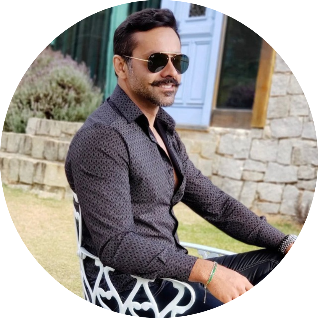Man with mustache and sunglasses sitting on a white metal chair outdoors near stone wall and greenery.