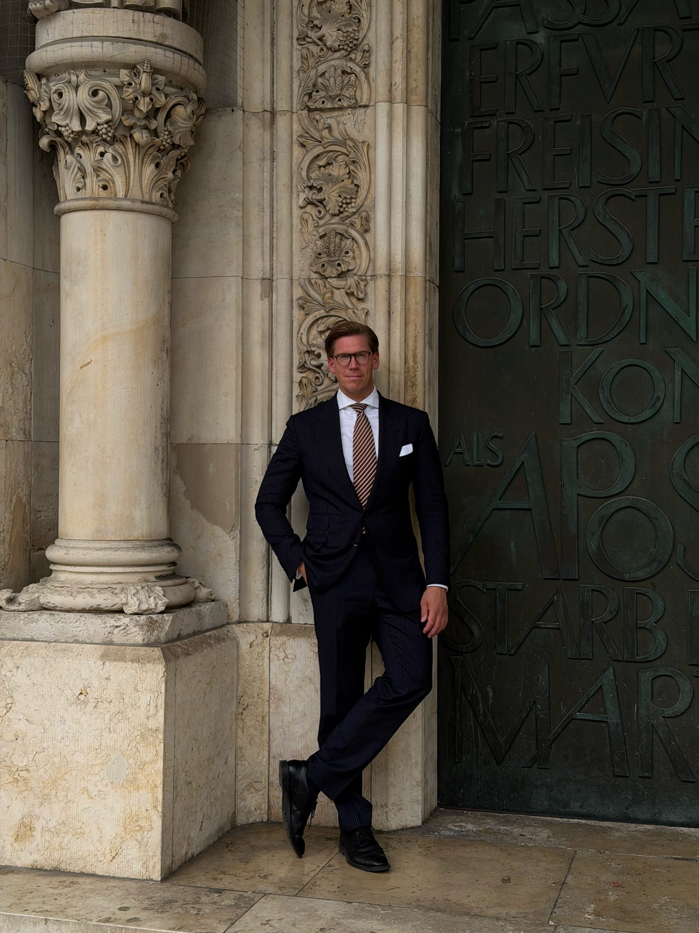 Man in a dark pinstripe suit, white shirt, striped tie, and glasses leaning against a textured stone wall next to a large ornate door with raised letters.