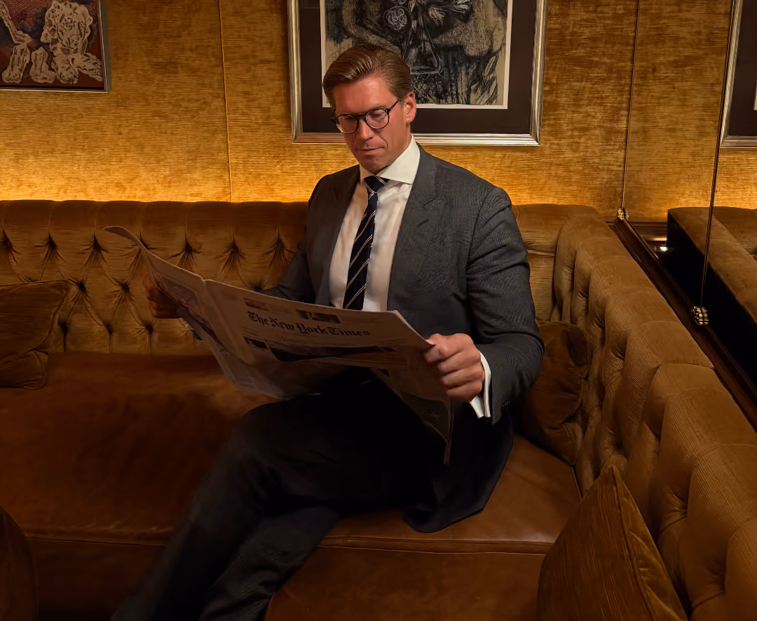 A man in a dark suit, white shirt, striped tie, and glasses sits on a tan tufted leather sofa, reading a newspaper with the headline The New York Times in a warmly lit room with framed artwork on the walls.