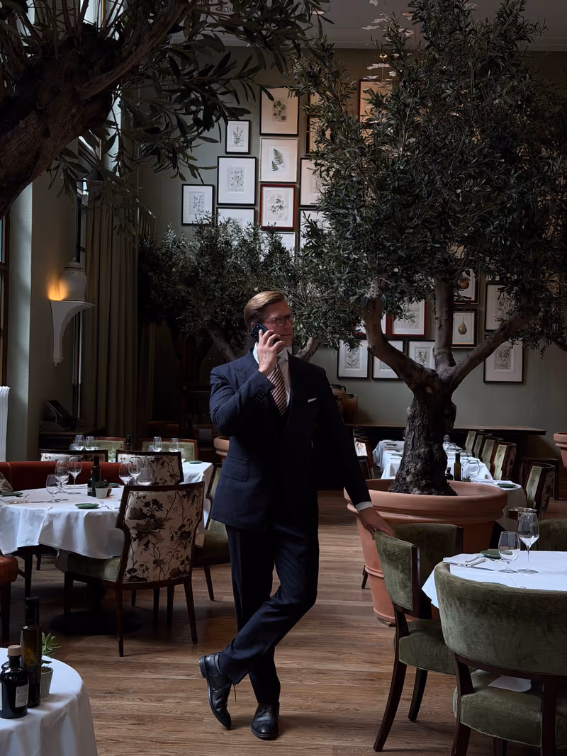 A man in a dark suit and striped tie talking on a cellphone while walking through an elegant restaurant with white tablecloths, green chairs, large potted trees, and framed botanical artwork on the walls.