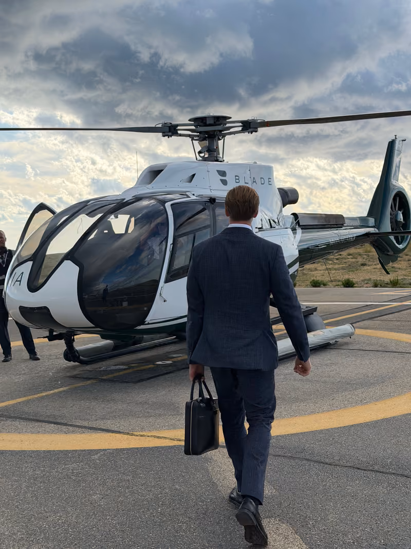 Man in a suit with a briefcase walking towards a white and black Blade helicopter on a helipad under a cloudy sky.