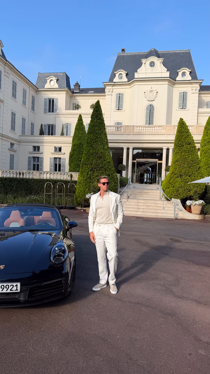 Man dressed in an all-white suit and sunglasses standing beside a black convertible Porsche with red interior in front of a large, elegant mansion with manicured tall triangular shrubs and a stairway entrance.