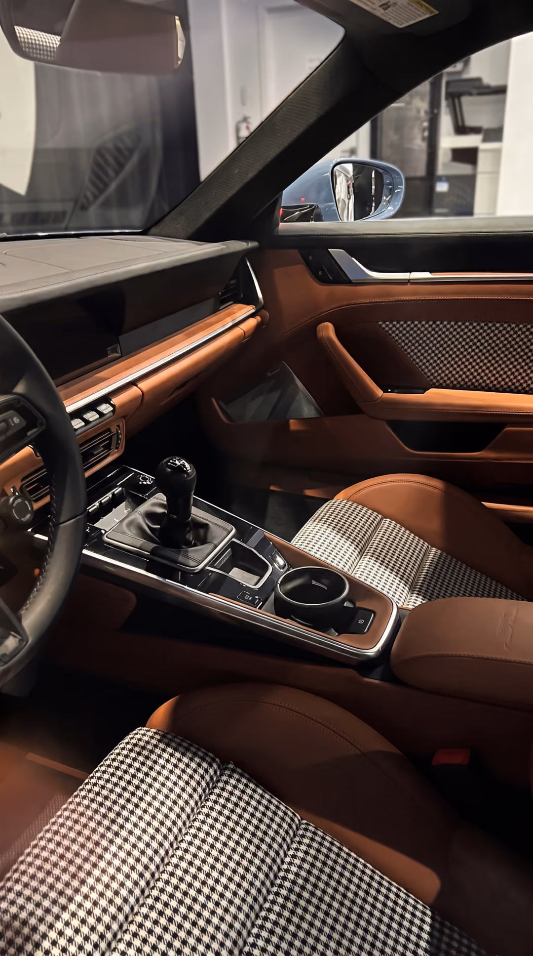 Interior of a car showing front seats with brown leather sides and black-and-white houndstooth fabric center, brown leather door panel, and a black manual gear shift on a center console with cup holder.