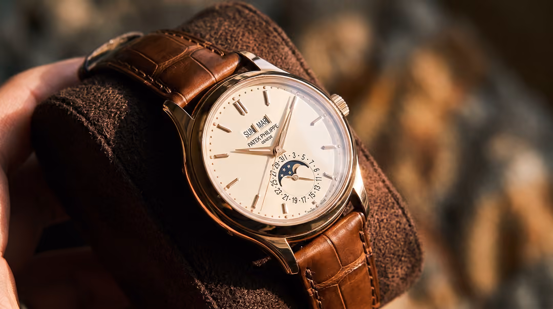 Close-up of a luxury Patek Philippe wristwatch with a brown leather strap, featuring a white dial, date and moon phase indicators, displayed on a brown cushion.