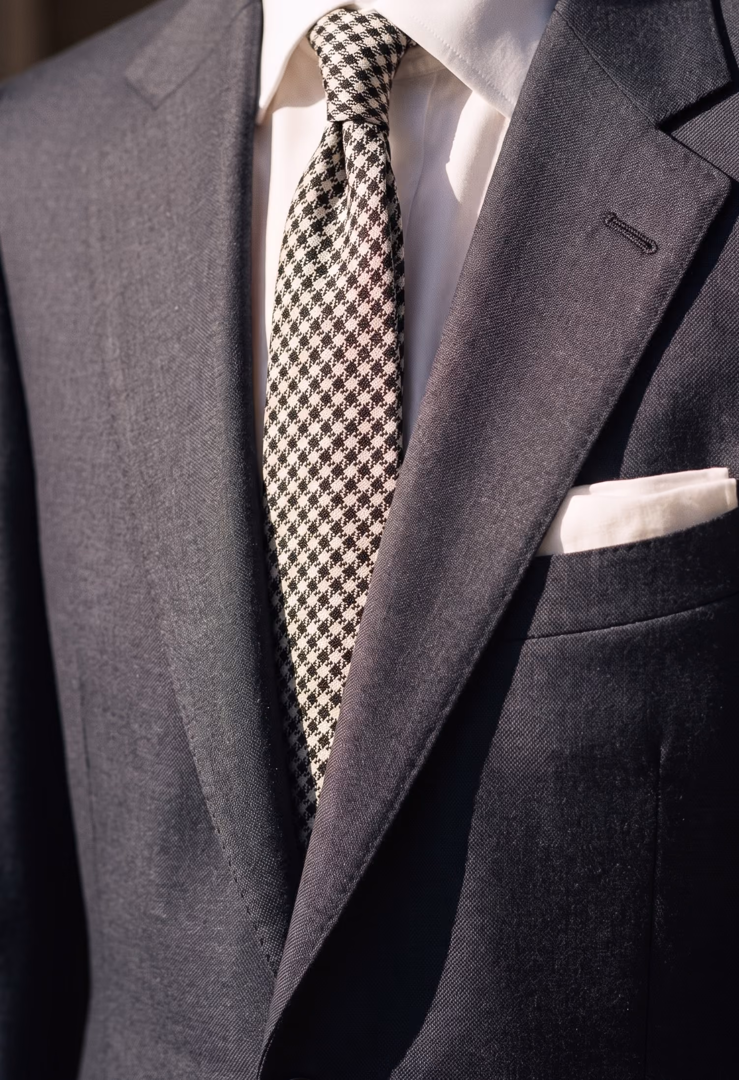 Close-up of a dark gray suit jacket over a white dress shirt, with a black and white houndstooth patterned tie and a white pocket square.