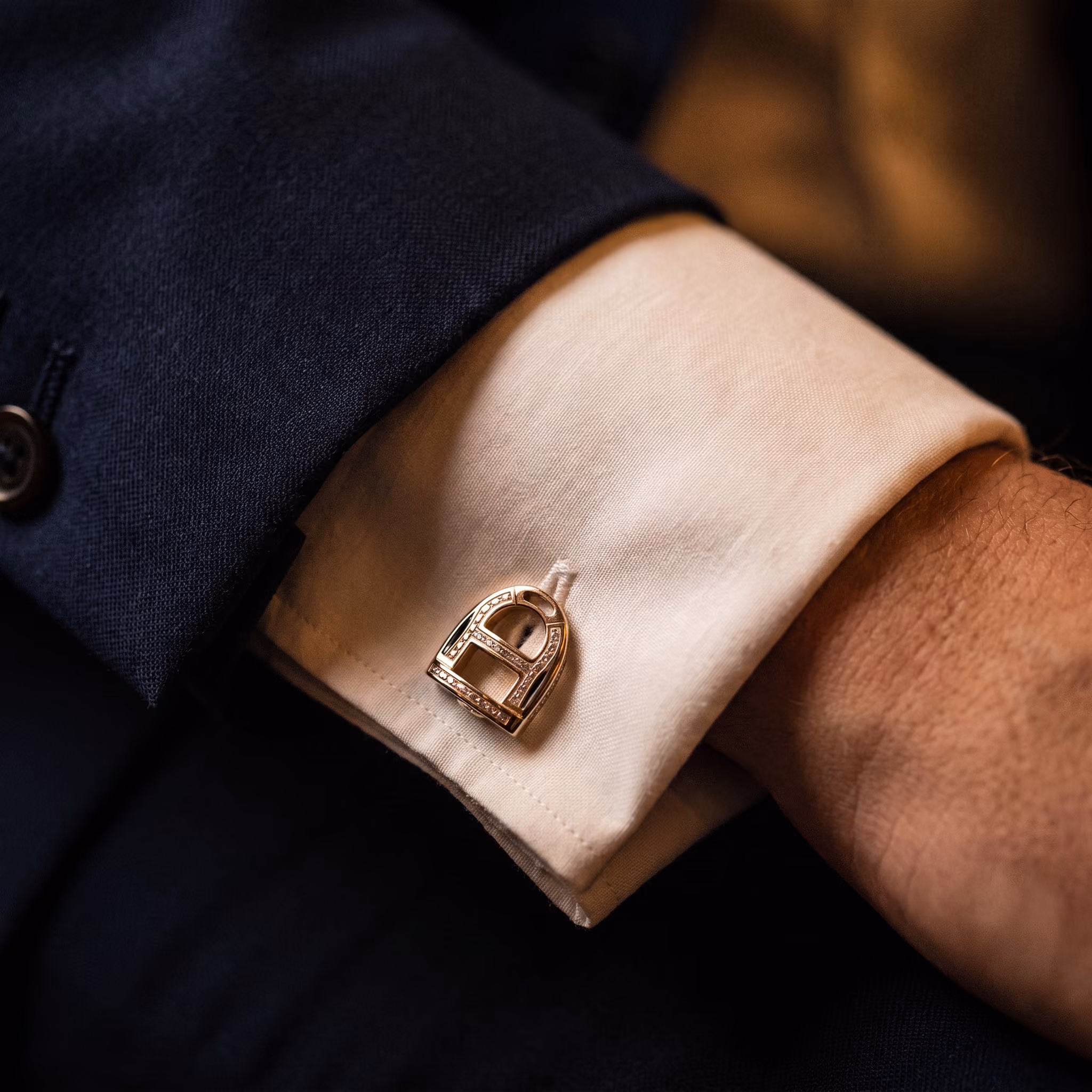 Close-up of a person's wrist wearing a white dress shirt with a gold and diamond-studded cufflink shaped like a stylized lock, partially covered by a dark suit sleeve.