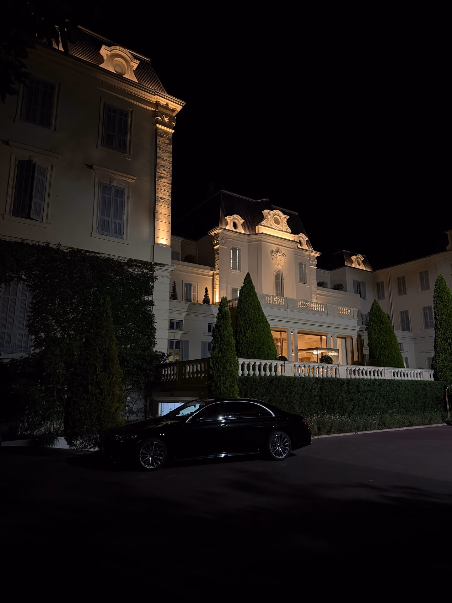 Night view of an elegant, illuminated mansion with tall trimmed trees and a black car parked in front.