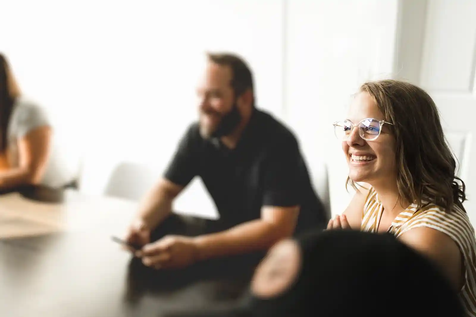 Smiling woman with glasses sitting at a table, alongside blurred people in a bright room.