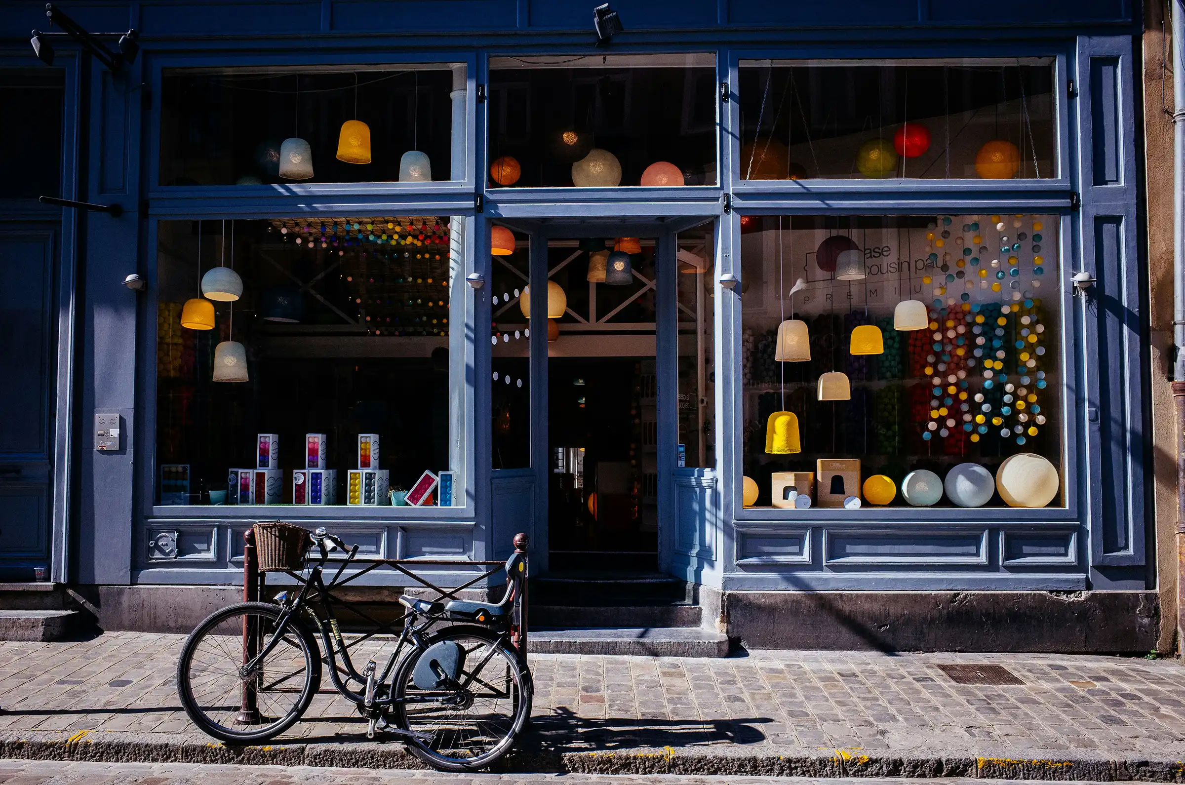 Black bicycle parked on cobblestone street in front of a blue shop window displaying hanging and table lamps in various colors and shapes.