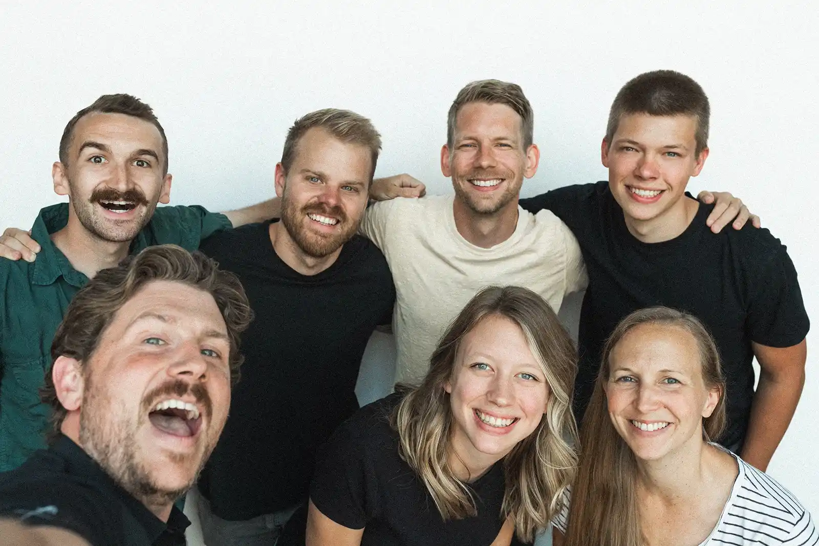 Group of seven smiling young adults, four men standing and three people sitting in front, taking a selfie against a plain white background.
