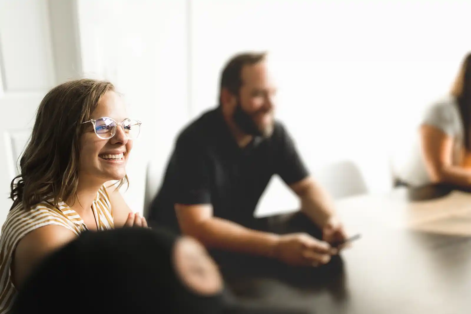 Smiling woman with glasses in striped top sitting at a table with blurred people in the background during a meeting.