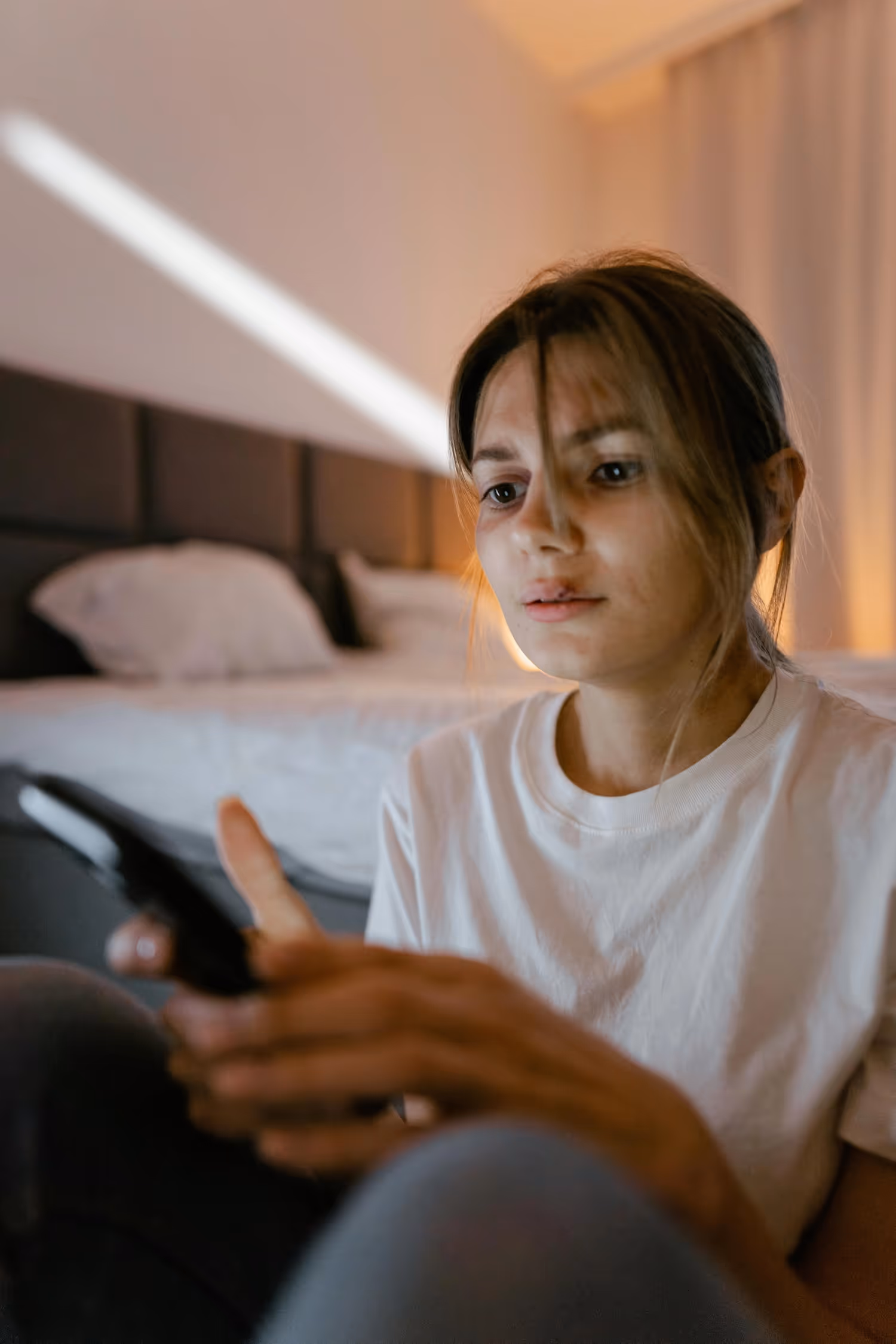 Young woman with bruises on her face sitting indoors, looking intently at her smartphone with a blurred bed in the background.