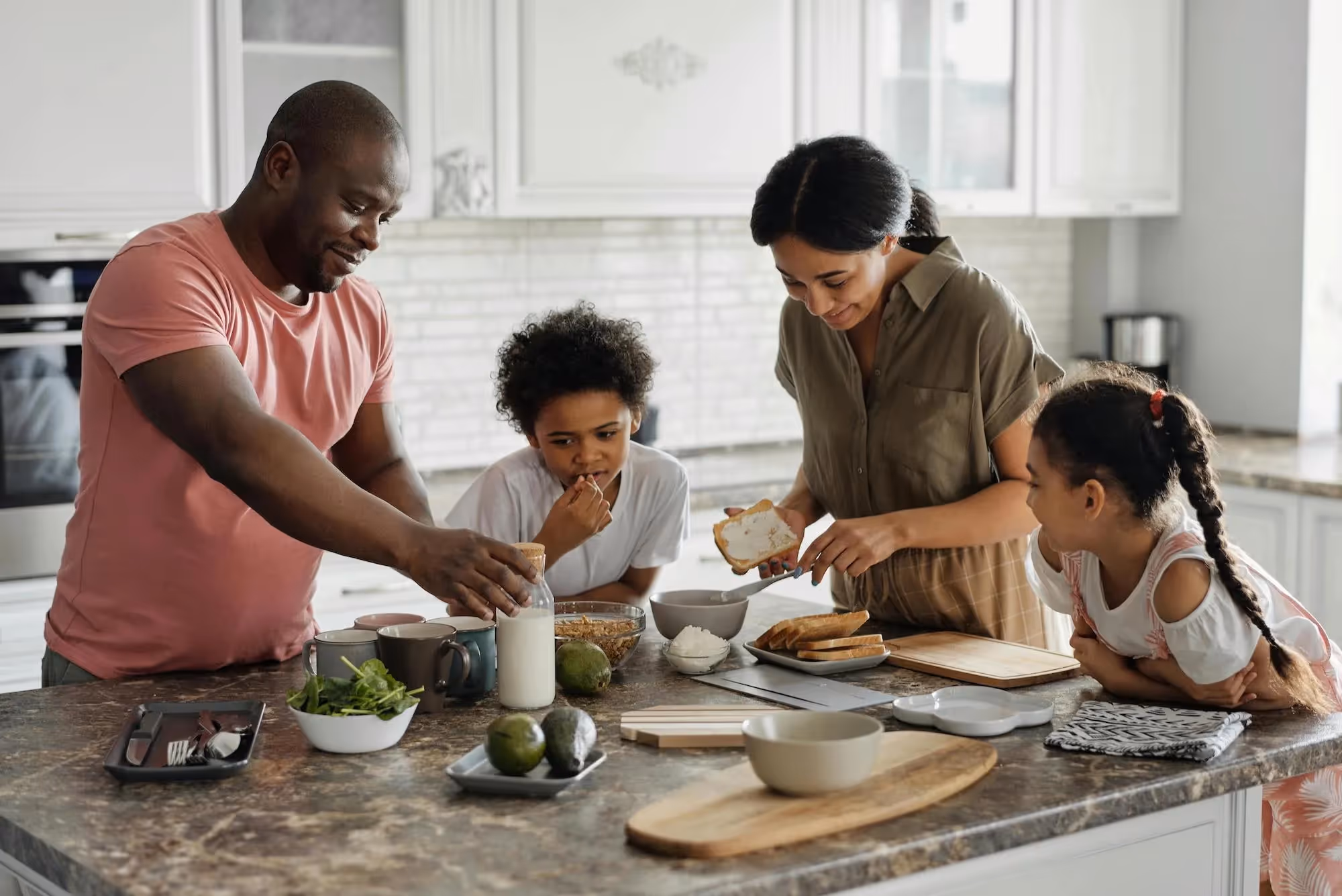 Family of four preparing breakfast together in a kitchen, with parents spreading cream cheese on bread and children watching.